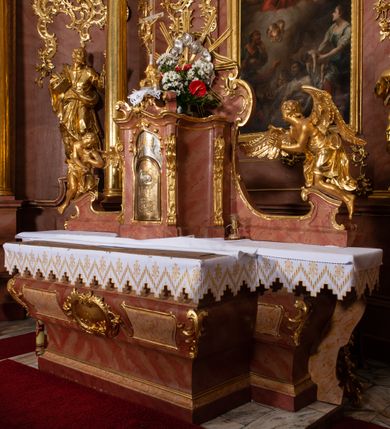 Zdjęcie nr 16: Nastawa ołtarzowa architektoniczna, jednokondygnacyjna ze zwieńczeniem, pięcioosiowa, ustawiona na wysokim cokole. Ołtarz wolnostojący, drewniany, sarkofagowy, wklęsło-wypukły, o zdwojonym froncie; mensa drewniana z marmurowym portatylem. Od frontu rocaille&#039;owy kartusz ujęty fantazyjnymi płycinami oraz liśćmi. Na ołtarzu tabernakulum w formie sześcianu o wklęsłych bokach i wklęsło-wypukłym froncie; ujęte w narożach dwiema parami pilastrów o trzonach przechodzących w spływy wolutowe zdobione rocaille&#039;m i różami. Pilastry podtrzymują falujący gzyms oraz fragmenty falistego, przerwanego, odwróconego przyczółka, przechodzącego w cokół, stanowiący tron wystawienia, na którym stoi krucyfiks. Na ściance frontowej obrotowa nisza, o wykroju stojącego prostokąta zamkniętego łukiem półkolistym, w opasce, zwieńczona kampanulami. W jej polu drzwiczki w formie stojącego prostokąta zamkniętego łukiem półkolistym w profilowanej ramie, z odlewanym przedstawieniem baranka leżącego na księdze z pieczęciami. Tabernakulum ujęte spływami przechodzącymi na krańcach we fragmenty wolutowego gzymsu, na których siedzą figury adorujących aniołów. Ponad tabernakulum fantazyjna ścianka tworząca tło dla tronu wystawienia z prześwitem otoczonym obłokami, z glorią promienistą, flankowana rocaille&#039;owymi uszami. Cokół nastawy z wyodrębnionymi dwoma prostopadłościanami i dwoma konsolami, na dwóch skrajnych osiach bramki zamknięte wolutowymi szczytami tworzącymi cokoły dla figur, z przejściami w formie stojącego prostokąta zamkniętego łukiem odcinkowym nadwieszonym, z drzwiczkami. Cokół i drzwiczki dekorowane rocaille&#039;m. Lico nastawy wklęsłe, osie wydzielone trzema parami pilastrów, na których tle stoją dwie pary kolumn o kapitelach kompozytowych. Podpory dźwigają pełne belkowanie, wyłamane na ich osi, przerwane nad polem środkowym, o gzymsie przechodzącym miejscami w rocaille&#039;owe kartusze. Na osi środkowej wnęka z obrazem Matki Boskiej Wspomożenia Wiernych, w formie stojącego prostokąta zamkniętego łukiem odcinkowym nadwieszonym z uskokiem, ujęta profilowaną ramą, dekorowaną ponad łukiem płomienistym rocaille&#039;m. Na pozostałych czterech osiach figury św. Barbary, św. Apolonii, św. Piotra i św. Pawła ustawione na rokokowych wspornikach. Trzony pilastrów i pola osi bocznych wypełnione rocaille&#039;m, fryz dekorowany zwisami kwiatowymi. Zwieńczenie w formie zbliżonej do trapezu, zamknięte dwoma fragmentami gzymsu przechodzącego w woluty, flankowane wolutami spływającymi na belkowanie wewnętrznej pary kolumn, na których siedzą adorujące figury aniołów. W polu rzeźbiarska grupa Trójcy Świętej, pośród obłoków, w glorii promienistej, zwieńczona gołębicą Ducha Świętego w otoku z obłoków i uskrzydlonych główek anielskich, w glorii promienistej, flankowana przez dwa putta siedzące na gzymsie. Zwieńczenie po bokach obwiedzione rocaille&#039;m, ujęte liśćmi palmowymi. Na belkowaniu, na osiach zewnętrznej pary kolumn wolutowe cokoły z rokokowymi wazonami. Struktura polichromowana na sposób imitujący marmoryzację w kolorze różowym i różowo-pomarańczowym, kolumny, profile i ornamentyka złocone, obłoki srebrzone. 

