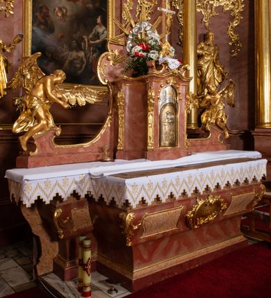 Zdjęcie nr 9: Nastawa ołtarzowa architektoniczna, jednokondygnacyjna ze zwieńczeniem, pięcioosiowa, ustawiona na wysokim cokole. Ołtarz wolnostojący, drewniany, sarkofagowy, wklęsło-wypukły, o zdwojonym froncie; mensa drewniana z marmurowym portatylem. Od frontu rocaille'owy kartusz ujęty fantazyjnymi płycinami oraz liśćmi. Na ołtarzu tabernakulum w formie sześcianu o wklęsłych bokach i wklęsło-wypukłym froncie; ujęte w narożach dwiema parami pilastrów o trzonach przechodzących w spływy wolutowe zdobione rocaille'm i różami. Pilastry podtrzymują falujący gzyms oraz fragmenty falistego, przerwanego, odwróconego przyczółka, przechodzącego w cokół, stanowiący tron wystawienia, na którym stoi krucyfiks. Na ściance frontowej obrotowa nisza, o wykroju stojącego prostokąta zamkniętego łukiem półkolistym, w opasce, zwieńczona kampanulami. W jej polu drzwiczki w formie stojącego prostokąta zamkniętego łukiem półkolistym w profilowanej ramie, z odlewanym przedstawieniem baranka leżącego na księdze z pieczęciami. Tabernakulum ujęte spływami przechodzącymi na krańcach we fragmenty wolutowego gzymsu, na których siedzą figury adorujących aniołów. Ponad tabernakulum fantazyjna ścianka tworząca tło dla tronu wystawienia z prześwitem otoczonym obłokami, z glorią promienistą, flankowana rocaille'owymi uszami. Cokół nastawy z wyodrębnionymi dwoma prostopadłościanami i dwoma konsolami, na dwóch skrajnych osiach bramki zamknięte wolutowymi szczytami tworzącymi cokoły dla figur, z przejściami w formie stojącego prostokąta zamkniętego łukiem odcinkowym nadwieszonym, z drzwiczkami. Cokół i drzwiczki dekorowane rocaille'm. Lico nastawy wklęsłe, osie wydzielone trzema parami pilastrów, na których tle stoją dwie pary kolumn o kapitelach kompozytowych. Podpory dźwigają pełne belkowanie, wyłamane na ich osi, przerwane nad polem środkowym, o gzymsie przechodzącym miejscami w rocaille'owe kartusze. Na osi środkowej wnęka z obrazem Matki Boskiej Wspomożenia Wiernych, w formie stojącego prostokąta zamkniętego łukiem odcinkowym nadwieszonym z uskokiem, ujęta profilowaną ramą, dekorowaną ponad łukiem płomienistym rocaille'm. Na pozostałych czterech osiach figury św. Barbary, św. Apolonii, św. Piotra i św. Pawła ustawione na rokokowych wspornikach. Trzony pilastrów i pola osi bocznych wypełnione rocaille'm, fryz dekorowany zwisami kwiatowymi. Zwieńczenie w formie zbliżonej do trapezu, zamknięte dwoma fragmentami gzymsu przechodzącego w woluty, flankowane wolutami spływającymi na belkowanie wewnętrznej pary kolumn, na których siedzą adorujące figury aniołów. W polu rzeźbiarska grupa Trójcy Świętej, pośród obłoków, w glorii promienistej, zwieńczona gołębicą Ducha Świętego w otoku z obłoków i uskrzydlonych główek anielskich, w glorii promienistej, flankowana przez dwa putta siedzące na gzymsie. Zwieńczenie po bokach obwiedzione rocaille'm, ujęte liśćmi palmowymi. Na belkowaniu, na osiach zewnętrznej pary kolumn wolutowe cokoły z rokokowymi wazonami. Struktura polichromowana na sposób imitujący marmoryzację w kolorze różowym i różowo-pomarańczowym, kolumny, profile i ornamentyka złocone, obłoki srebrzone. 

