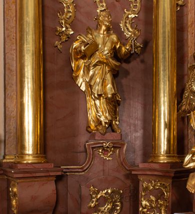 Zdjęcie nr 19: Nastawa ołtarzowa architektoniczna, jednokondygnacyjna ze zwieńczeniem, pięcioosiowa, ustawiona na wysokim cokole. Ołtarz wolnostojący, drewniany, sarkofagowy, wklęsło-wypukły, o zdwojonym froncie; mensa drewniana z marmurowym portatylem. Od frontu rocaille'owy kartusz ujęty fantazyjnymi płycinami oraz liśćmi. Na ołtarzu tabernakulum w formie sześcianu o wklęsłych bokach i wklęsło-wypukłym froncie; ujęte w narożach dwiema parami pilastrów o trzonach przechodzących w spływy wolutowe zdobione rocaille'm i różami. Pilastry podtrzymują falujący gzyms oraz fragmenty falistego, przerwanego, odwróconego przyczółka, przechodzącego w cokół, stanowiący tron wystawienia, na którym stoi krucyfiks. Na ściance frontowej obrotowa nisza, o wykroju stojącego prostokąta zamkniętego łukiem półkolistym, w opasce, zwieńczona kampanulami. W jej polu drzwiczki w formie stojącego prostokąta zamkniętego łukiem półkolistym w profilowanej ramie, z odlewanym przedstawieniem baranka leżącego na księdze z pieczęciami. Tabernakulum ujęte spływami przechodzącymi na krańcach we fragmenty wolutowego gzymsu, na których siedzą figury adorujących aniołów. Ponad tabernakulum fantazyjna ścianka tworząca tło dla tronu wystawienia z prześwitem otoczonym obłokami, z glorią promienistą, flankowana rocaille'owymi uszami. Cokół nastawy z wyodrębnionymi dwoma prostopadłościanami i dwoma konsolami, na dwóch skrajnych osiach bramki zamknięte wolutowymi szczytami tworzącymi cokoły dla figur, z przejściami w formie stojącego prostokąta zamkniętego łukiem odcinkowym nadwieszonym, z drzwiczkami. Cokół i drzwiczki dekorowane rocaille'm. Lico nastawy wklęsłe, osie wydzielone trzema parami pilastrów, na których tle stoją dwie pary kolumn o kapitelach kompozytowych. Podpory dźwigają pełne belkowanie, wyłamane na ich osi, przerwane nad polem środkowym, o gzymsie przechodzącym miejscami w rocaille'owe kartusze. Na osi środkowej wnęka z obrazem Matki Boskiej Wspomożenia Wiernych, w formie stojącego prostokąta zamkniętego łukiem odcinkowym nadwieszonym z uskokiem, ujęta profilowaną ramą, dekorowaną ponad łukiem płomienistym rocaille'm. Na pozostałych czterech osiach figury św. Barbary, św. Apolonii, św. Piotra i św. Pawła ustawione na rokokowych wspornikach. Trzony pilastrów i pola osi bocznych wypełnione rocaille'm, fryz dekorowany zwisami kwiatowymi. Zwieńczenie w formie zbliżonej do trapezu, zamknięte dwoma fragmentami gzymsu przechodzącego w woluty, flankowane wolutami spływającymi na belkowanie wewnętrznej pary kolumn, na których siedzą adorujące figury aniołów. W polu rzeźbiarska grupa Trójcy Świętej, pośród obłoków, w glorii promienistej, zwieńczona gołębicą Ducha Świętego w otoku z obłoków i uskrzydlonych główek anielskich, w glorii promienistej, flankowana przez dwa putta siedzące na gzymsie. Zwieńczenie po bokach obwiedzione rocaille'm, ujęte liśćmi palmowymi. Na belkowaniu, na osiach zewnętrznej pary kolumn wolutowe cokoły z rokokowymi wazonami. Struktura polichromowana na sposób imitujący marmoryzację w kolorze różowym i różowo-pomarańczowym, kolumny, profile i ornamentyka złocone, obłoki srebrzone. 

