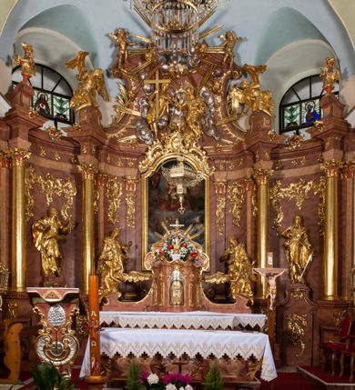 Zdjęcie nr 7: Nastawa ołtarzowa architektoniczna, jednokondygnacyjna ze zwieńczeniem, pięcioosiowa, ustawiona na wysokim cokole. Ołtarz wolnostojący, drewniany, sarkofagowy, wklęsło-wypukły, o zdwojonym froncie; mensa drewniana z marmurowym portatylem. Od frontu rocaille'owy kartusz ujęty fantazyjnymi płycinami oraz liśćmi. Na ołtarzu tabernakulum w formie sześcianu o wklęsłych bokach i wklęsło-wypukłym froncie; ujęte w narożach dwiema parami pilastrów o trzonach przechodzących w spływy wolutowe zdobione rocaille'm i różami. Pilastry podtrzymują falujący gzyms oraz fragmenty falistego, przerwanego, odwróconego przyczółka, przechodzącego w cokół, stanowiący tron wystawienia, na którym stoi krucyfiks. Na ściance frontowej obrotowa nisza, o wykroju stojącego prostokąta zamkniętego łukiem półkolistym, w opasce, zwieńczona kampanulami. W jej polu drzwiczki w formie stojącego prostokąta zamkniętego łukiem półkolistym w profilowanej ramie, z odlewanym przedstawieniem baranka leżącego na księdze z pieczęciami. Tabernakulum ujęte spływami przechodzącymi na krańcach we fragmenty wolutowego gzymsu, na których siedzą figury adorujących aniołów. Ponad tabernakulum fantazyjna ścianka tworząca tło dla tronu wystawienia z prześwitem otoczonym obłokami, z glorią promienistą, flankowana rocaille'owymi uszami. Cokół nastawy z wyodrębnionymi dwoma prostopadłościanami i dwoma konsolami, na dwóch skrajnych osiach bramki zamknięte wolutowymi szczytami tworzącymi cokoły dla figur, z przejściami w formie stojącego prostokąta zamkniętego łukiem odcinkowym nadwieszonym, z drzwiczkami. Cokół i drzwiczki dekorowane rocaille'm. Lico nastawy wklęsłe, osie wydzielone trzema parami pilastrów, na których tle stoją dwie pary kolumn o kapitelach kompozytowych. Podpory dźwigają pełne belkowanie, wyłamane na ich osi, przerwane nad polem środkowym, o gzymsie przechodzącym miejscami w rocaille'owe kartusze. Na osi środkowej wnęka z obrazem Matki Boskiej Wspomożenia Wiernych, w formie stojącego prostokąta zamkniętego łukiem odcinkowym nadwieszonym z uskokiem, ujęta profilowaną ramą, dekorowaną ponad łukiem płomienistym rocaille'm. Na pozostałych czterech osiach figury św. Barbary, św. Apolonii, św. Piotra i św. Pawła ustawione na rokokowych wspornikach. Trzony pilastrów i pola osi bocznych wypełnione rocaille'm, fryz dekorowany zwisami kwiatowymi. Zwieńczenie w formie zbliżonej do trapezu, zamknięte dwoma fragmentami gzymsu przechodzącego w woluty, flankowane wolutami spływającymi na belkowanie wewnętrznej pary kolumn, na których siedzą adorujące figury aniołów. W polu rzeźbiarska grupa Trójcy Świętej, pośród obłoków, w glorii promienistej, zwieńczona gołębicą Ducha Świętego w otoku z obłoków i uskrzydlonych główek anielskich, w glorii promienistej, flankowana przez dwa putta siedzące na gzymsie. Zwieńczenie po bokach obwiedzione rocaille'm, ujęte liśćmi palmowymi. Na belkowaniu, na osiach zewnętrznej pary kolumn wolutowe cokoły z rokokowymi wazonami. Struktura polichromowana na sposób imitujący marmoryzację w kolorze różowym i różowo-pomarańczowym, kolumny, profile i ornamentyka złocone, obłoki srebrzone. 

