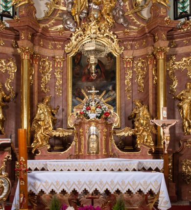 Zdjęcie nr 8: Nastawa ołtarzowa architektoniczna, jednokondygnacyjna ze zwieńczeniem, pięcioosiowa, ustawiona na wysokim cokole. Ołtarz wolnostojący, drewniany, sarkofagowy, wklęsło-wypukły, o zdwojonym froncie; mensa drewniana z marmurowym portatylem. Od frontu rocaille'owy kartusz ujęty fantazyjnymi płycinami oraz liśćmi. Na ołtarzu tabernakulum w formie sześcianu o wklęsłych bokach i wklęsło-wypukłym froncie; ujęte w narożach dwiema parami pilastrów o trzonach przechodzących w spływy wolutowe zdobione rocaille'm i różami. Pilastry podtrzymują falujący gzyms oraz fragmenty falistego, przerwanego, odwróconego przyczółka, przechodzącego w cokół, stanowiący tron wystawienia, na którym stoi krucyfiks. Na ściance frontowej obrotowa nisza, o wykroju stojącego prostokąta zamkniętego łukiem półkolistym, w opasce, zwieńczona kampanulami. W jej polu drzwiczki w formie stojącego prostokąta zamkniętego łukiem półkolistym w profilowanej ramie, z odlewanym przedstawieniem baranka leżącego na księdze z pieczęciami. Tabernakulum ujęte spływami przechodzącymi na krańcach we fragmenty wolutowego gzymsu, na których siedzą figury adorujących aniołów. Ponad tabernakulum fantazyjna ścianka tworząca tło dla tronu wystawienia z prześwitem otoczonym obłokami, z glorią promienistą, flankowana rocaille'owymi uszami. Cokół nastawy z wyodrębnionymi dwoma prostopadłościanami i dwoma konsolami, na dwóch skrajnych osiach bramki zamknięte wolutowymi szczytami tworzącymi cokoły dla figur, z przejściami w formie stojącego prostokąta zamkniętego łukiem odcinkowym nadwieszonym, z drzwiczkami. Cokół i drzwiczki dekorowane rocaille'm. Lico nastawy wklęsłe, osie wydzielone trzema parami pilastrów, na których tle stoją dwie pary kolumn o kapitelach kompozytowych. Podpory dźwigają pełne belkowanie, wyłamane na ich osi, przerwane nad polem środkowym, o gzymsie przechodzącym miejscami w rocaille'owe kartusze. Na osi środkowej wnęka z obrazem Matki Boskiej Wspomożenia Wiernych, w formie stojącego prostokąta zamkniętego łukiem odcinkowym nadwieszonym z uskokiem, ujęta profilowaną ramą, dekorowaną ponad łukiem płomienistym rocaille'm. Na pozostałych czterech osiach figury św. Barbary, św. Apolonii, św. Piotra i św. Pawła ustawione na rokokowych wspornikach. Trzony pilastrów i pola osi bocznych wypełnione rocaille'm, fryz dekorowany zwisami kwiatowymi. Zwieńczenie w formie zbliżonej do trapezu, zamknięte dwoma fragmentami gzymsu przechodzącego w woluty, flankowane wolutami spływającymi na belkowanie wewnętrznej pary kolumn, na których siedzą adorujące figury aniołów. W polu rzeźbiarska grupa Trójcy Świętej, pośród obłoków, w glorii promienistej, zwieńczona gołębicą Ducha Świętego w otoku z obłoków i uskrzydlonych główek anielskich, w glorii promienistej, flankowana przez dwa putta siedzące na gzymsie. Zwieńczenie po bokach obwiedzione rocaille'm, ujęte liśćmi palmowymi. Na belkowaniu, na osiach zewnętrznej pary kolumn wolutowe cokoły z rokokowymi wazonami. Struktura polichromowana na sposób imitujący marmoryzację w kolorze różowym i różowo-pomarańczowym, kolumny, profile i ornamentyka złocone, obłoki srebrzone. 

