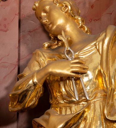 Zdjęcie nr 21: Nastawa ołtarzowa architektoniczna, jednokondygnacyjna ze zwieńczeniem, pięcioosiowa, ustawiona na wysokim cokole. Ołtarz wolnostojący, drewniany, sarkofagowy, wklęsło-wypukły, o zdwojonym froncie; mensa drewniana z marmurowym portatylem. Od frontu rocaille&#039;owy kartusz ujęty fantazyjnymi płycinami oraz liśćmi. Na ołtarzu tabernakulum w formie sześcianu o wklęsłych bokach i wklęsło-wypukłym froncie; ujęte w narożach dwiema parami pilastrów o trzonach przechodzących w spływy wolutowe zdobione rocaille&#039;m i różami. Pilastry podtrzymują falujący gzyms oraz fragmenty falistego, przerwanego, odwróconego przyczółka, przechodzącego w cokół, stanowiący tron wystawienia, na którym stoi krucyfiks. Na ściance frontowej obrotowa nisza, o wykroju stojącego prostokąta zamkniętego łukiem półkolistym, w opasce, zwieńczona kampanulami. W jej polu drzwiczki w formie stojącego prostokąta zamkniętego łukiem półkolistym w profilowanej ramie, z odlewanym przedstawieniem baranka leżącego na księdze z pieczęciami. Tabernakulum ujęte spływami przechodzącymi na krańcach we fragmenty wolutowego gzymsu, na których siedzą figury adorujących aniołów. Ponad tabernakulum fantazyjna ścianka tworząca tło dla tronu wystawienia z prześwitem otoczonym obłokami, z glorią promienistą, flankowana rocaille&#039;owymi uszami. Cokół nastawy z wyodrębnionymi dwoma prostopadłościanami i dwoma konsolami, na dwóch skrajnych osiach bramki zamknięte wolutowymi szczytami tworzącymi cokoły dla figur, z przejściami w formie stojącego prostokąta zamkniętego łukiem odcinkowym nadwieszonym, z drzwiczkami. Cokół i drzwiczki dekorowane rocaille&#039;m. Lico nastawy wklęsłe, osie wydzielone trzema parami pilastrów, na których tle stoją dwie pary kolumn o kapitelach kompozytowych. Podpory dźwigają pełne belkowanie, wyłamane na ich osi, przerwane nad polem środkowym, o gzymsie przechodzącym miejscami w rocaille&#039;owe kartusze. Na osi środkowej wnęka z obrazem Matki Boskiej Wspomożenia Wiernych, w formie stojącego prostokąta zamkniętego łukiem odcinkowym nadwieszonym z uskokiem, ujęta profilowaną ramą, dekorowaną ponad łukiem płomienistym rocaille&#039;m. Na pozostałych czterech osiach figury św. Barbary, św. Apolonii, św. Piotra i św. Pawła ustawione na rokokowych wspornikach. Trzony pilastrów i pola osi bocznych wypełnione rocaille&#039;m, fryz dekorowany zwisami kwiatowymi. Zwieńczenie w formie zbliżonej do trapezu, zamknięte dwoma fragmentami gzymsu przechodzącego w woluty, flankowane wolutami spływającymi na belkowanie wewnętrznej pary kolumn, na których siedzą adorujące figury aniołów. W polu rzeźbiarska grupa Trójcy Świętej, pośród obłoków, w glorii promienistej, zwieńczona gołębicą Ducha Świętego w otoku z obłoków i uskrzydlonych główek anielskich, w glorii promienistej, flankowana przez dwa putta siedzące na gzymsie. Zwieńczenie po bokach obwiedzione rocaille&#039;m, ujęte liśćmi palmowymi. Na belkowaniu, na osiach zewnętrznej pary kolumn wolutowe cokoły z rokokowymi wazonami. Struktura polichromowana na sposób imitujący marmoryzację w kolorze różowym i różowo-pomarańczowym, kolumny, profile i ornamentyka złocone, obłoki srebrzone. 

