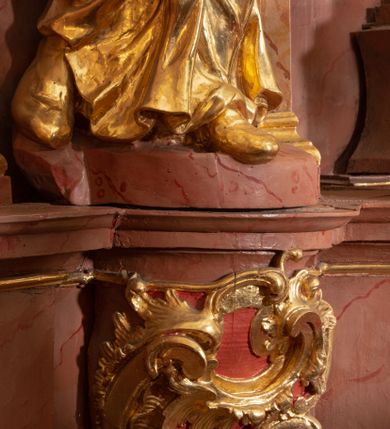 Zdjęcie nr 20: Nastawa ołtarzowa architektoniczna, jednokondygnacyjna ze zwieńczeniem, pięcioosiowa, ustawiona na wysokim cokole. Ołtarz wolnostojący, drewniany, sarkofagowy, wklęsło-wypukły, o zdwojonym froncie; mensa drewniana z marmurowym portatylem. Od frontu rocaille&#039;owy kartusz ujęty fantazyjnymi płycinami oraz liśćmi. Na ołtarzu tabernakulum w formie sześcianu o wklęsłych bokach i wklęsło-wypukłym froncie; ujęte w narożach dwiema parami pilastrów o trzonach przechodzących w spływy wolutowe zdobione rocaille&#039;m i różami. Pilastry podtrzymują falujący gzyms oraz fragmenty falistego, przerwanego, odwróconego przyczółka, przechodzącego w cokół, stanowiący tron wystawienia, na którym stoi krucyfiks. Na ściance frontowej obrotowa nisza, o wykroju stojącego prostokąta zamkniętego łukiem półkolistym, w opasce, zwieńczona kampanulami. W jej polu drzwiczki w formie stojącego prostokąta zamkniętego łukiem półkolistym w profilowanej ramie, z odlewanym przedstawieniem baranka leżącego na księdze z pieczęciami. Tabernakulum ujęte spływami przechodzącymi na krańcach we fragmenty wolutowego gzymsu, na których siedzą figury adorujących aniołów. Ponad tabernakulum fantazyjna ścianka tworząca tło dla tronu wystawienia z prześwitem otoczonym obłokami, z glorią promienistą, flankowana rocaille&#039;owymi uszami. Cokół nastawy z wyodrębnionymi dwoma prostopadłościanami i dwoma konsolami, na dwóch skrajnych osiach bramki zamknięte wolutowymi szczytami tworzącymi cokoły dla figur, z przejściami w formie stojącego prostokąta zamkniętego łukiem odcinkowym nadwieszonym, z drzwiczkami. Cokół i drzwiczki dekorowane rocaille&#039;m. Lico nastawy wklęsłe, osie wydzielone trzema parami pilastrów, na których tle stoją dwie pary kolumn o kapitelach kompozytowych. Podpory dźwigają pełne belkowanie, wyłamane na ich osi, przerwane nad polem środkowym, o gzymsie przechodzącym miejscami w rocaille&#039;owe kartusze. Na osi środkowej wnęka z obrazem Matki Boskiej Wspomożenia Wiernych, w formie stojącego prostokąta zamkniętego łukiem odcinkowym nadwieszonym z uskokiem, ujęta profilowaną ramą, dekorowaną ponad łukiem płomienistym rocaille&#039;m. Na pozostałych czterech osiach figury św. Barbary, św. Apolonii, św. Piotra i św. Pawła ustawione na rokokowych wspornikach. Trzony pilastrów i pola osi bocznych wypełnione rocaille&#039;m, fryz dekorowany zwisami kwiatowymi. Zwieńczenie w formie zbliżonej do trapezu, zamknięte dwoma fragmentami gzymsu przechodzącego w woluty, flankowane wolutami spływającymi na belkowanie wewnętrznej pary kolumn, na których siedzą adorujące figury aniołów. W polu rzeźbiarska grupa Trójcy Świętej, pośród obłoków, w glorii promienistej, zwieńczona gołębicą Ducha Świętego w otoku z obłoków i uskrzydlonych główek anielskich, w glorii promienistej, flankowana przez dwa putta siedzące na gzymsie. Zwieńczenie po bokach obwiedzione rocaille&#039;m, ujęte liśćmi palmowymi. Na belkowaniu, na osiach zewnętrznej pary kolumn wolutowe cokoły z rokokowymi wazonami. Struktura polichromowana na sposób imitujący marmoryzację w kolorze różowym i różowo-pomarańczowym, kolumny, profile i ornamentyka złocone, obłoki srebrzone. 

