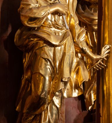 Zdjęcie nr 25: Nastawa ołtarzowa architektoniczna, jednokondygnacyjna ze zwieńczeniem, pięcioosiowa, ustawiona na wysokim cokole. Ołtarz wolnostojący, drewniany, sarkofagowy, wklęsło-wypukły, o zdwojonym froncie; mensa drewniana z marmurowym portatylem. Od frontu rocaille'owy kartusz ujęty fantazyjnymi płycinami oraz liśćmi. Na ołtarzu tabernakulum w formie sześcianu o wklęsłych bokach i wklęsło-wypukłym froncie; ujęte w narożach dwiema parami pilastrów o trzonach przechodzących w spływy wolutowe zdobione rocaille'm i różami. Pilastry podtrzymują falujący gzyms oraz fragmenty falistego, przerwanego, odwróconego przyczółka, przechodzącego w cokół, stanowiący tron wystawienia, na którym stoi krucyfiks. Na ściance frontowej obrotowa nisza, o wykroju stojącego prostokąta zamkniętego łukiem półkolistym, w opasce, zwieńczona kampanulami. W jej polu drzwiczki w formie stojącego prostokąta zamkniętego łukiem półkolistym w profilowanej ramie, z odlewanym przedstawieniem baranka leżącego na księdze z pieczęciami. Tabernakulum ujęte spływami przechodzącymi na krańcach we fragmenty wolutowego gzymsu, na których siedzą figury adorujących aniołów. Ponad tabernakulum fantazyjna ścianka tworząca tło dla tronu wystawienia z prześwitem otoczonym obłokami, z glorią promienistą, flankowana rocaille'owymi uszami. Cokół nastawy z wyodrębnionymi dwoma prostopadłościanami i dwoma konsolami, na dwóch skrajnych osiach bramki zamknięte wolutowymi szczytami tworzącymi cokoły dla figur, z przejściami w formie stojącego prostokąta zamkniętego łukiem odcinkowym nadwieszonym, z drzwiczkami. Cokół i drzwiczki dekorowane rocaille'm. Lico nastawy wklęsłe, osie wydzielone trzema parami pilastrów, na których tle stoją dwie pary kolumn o kapitelach kompozytowych. Podpory dźwigają pełne belkowanie, wyłamane na ich osi, przerwane nad polem środkowym, o gzymsie przechodzącym miejscami w rocaille'owe kartusze. Na osi środkowej wnęka z obrazem Matki Boskiej Wspomożenia Wiernych, w formie stojącego prostokąta zamkniętego łukiem odcinkowym nadwieszonym z uskokiem, ujęta profilowaną ramą, dekorowaną ponad łukiem płomienistym rocaille'm. Na pozostałych czterech osiach figury św. Barbary, św. Apolonii, św. Piotra i św. Pawła ustawione na rokokowych wspornikach. Trzony pilastrów i pola osi bocznych wypełnione rocaille'm, fryz dekorowany zwisami kwiatowymi. Zwieńczenie w formie zbliżonej do trapezu, zamknięte dwoma fragmentami gzymsu przechodzącego w woluty, flankowane wolutami spływającymi na belkowanie wewnętrznej pary kolumn, na których siedzą adorujące figury aniołów. W polu rzeźbiarska grupa Trójcy Świętej, pośród obłoków, w glorii promienistej, zwieńczona gołębicą Ducha Świętego w otoku z obłoków i uskrzydlonych główek anielskich, w glorii promienistej, flankowana przez dwa putta siedzące na gzymsie. Zwieńczenie po bokach obwiedzione rocaille'm, ujęte liśćmi palmowymi. Na belkowaniu, na osiach zewnętrznej pary kolumn wolutowe cokoły z rokokowymi wazonami. Struktura polichromowana na sposób imitujący marmoryzację w kolorze różowym i różowo-pomarańczowym, kolumny, profile i ornamentyka złocone, obłoki srebrzone. 

