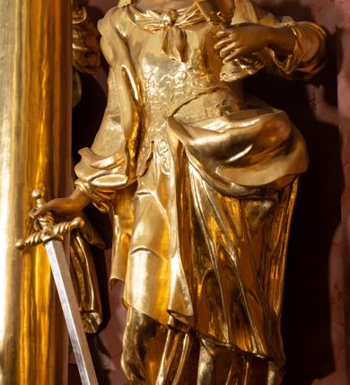 Zdjęcie nr 26: Nastawa ołtarzowa architektoniczna, jednokondygnacyjna ze zwieńczeniem, pięcioosiowa, ustawiona na wysokim cokole. Ołtarz wolnostojący, drewniany, sarkofagowy, wklęsło-wypukły, o zdwojonym froncie; mensa drewniana z marmurowym portatylem. Od frontu rocaille'owy kartusz ujęty fantazyjnymi płycinami oraz liśćmi. Na ołtarzu tabernakulum w formie sześcianu o wklęsłych bokach i wklęsło-wypukłym froncie; ujęte w narożach dwiema parami pilastrów o trzonach przechodzących w spływy wolutowe zdobione rocaille'm i różami. Pilastry podtrzymują falujący gzyms oraz fragmenty falistego, przerwanego, odwróconego przyczółka, przechodzącego w cokół, stanowiący tron wystawienia, na którym stoi krucyfiks. Na ściance frontowej obrotowa nisza, o wykroju stojącego prostokąta zamkniętego łukiem półkolistym, w opasce, zwieńczona kampanulami. W jej polu drzwiczki w formie stojącego prostokąta zamkniętego łukiem półkolistym w profilowanej ramie, z odlewanym przedstawieniem baranka leżącego na księdze z pieczęciami. Tabernakulum ujęte spływami przechodzącymi na krańcach we fragmenty wolutowego gzymsu, na których siedzą figury adorujących aniołów. Ponad tabernakulum fantazyjna ścianka tworząca tło dla tronu wystawienia z prześwitem otoczonym obłokami, z glorią promienistą, flankowana rocaille'owymi uszami. Cokół nastawy z wyodrębnionymi dwoma prostopadłościanami i dwoma konsolami, na dwóch skrajnych osiach bramki zamknięte wolutowymi szczytami tworzącymi cokoły dla figur, z przejściami w formie stojącego prostokąta zamkniętego łukiem odcinkowym nadwieszonym, z drzwiczkami. Cokół i drzwiczki dekorowane rocaille'm. Lico nastawy wklęsłe, osie wydzielone trzema parami pilastrów, na których tle stoją dwie pary kolumn o kapitelach kompozytowych. Podpory dźwigają pełne belkowanie, wyłamane na ich osi, przerwane nad polem środkowym, o gzymsie przechodzącym miejscami w rocaille'owe kartusze. Na osi środkowej wnęka z obrazem Matki Boskiej Wspomożenia Wiernych, w formie stojącego prostokąta zamkniętego łukiem odcinkowym nadwieszonym z uskokiem, ujęta profilowaną ramą, dekorowaną ponad łukiem płomienistym rocaille'm. Na pozostałych czterech osiach figury św. Barbary, św. Apolonii, św. Piotra i św. Pawła ustawione na rokokowych wspornikach. Trzony pilastrów i pola osi bocznych wypełnione rocaille'm, fryz dekorowany zwisami kwiatowymi. Zwieńczenie w formie zbliżonej do trapezu, zamknięte dwoma fragmentami gzymsu przechodzącego w woluty, flankowane wolutami spływającymi na belkowanie wewnętrznej pary kolumn, na których siedzą adorujące figury aniołów. W polu rzeźbiarska grupa Trójcy Świętej, pośród obłoków, w glorii promienistej, zwieńczona gołębicą Ducha Świętego w otoku z obłoków i uskrzydlonych główek anielskich, w glorii promienistej, flankowana przez dwa putta siedzące na gzymsie. Zwieńczenie po bokach obwiedzione rocaille'm, ujęte liśćmi palmowymi. Na belkowaniu, na osiach zewnętrznej pary kolumn wolutowe cokoły z rokokowymi wazonami. Struktura polichromowana na sposób imitujący marmoryzację w kolorze różowym i różowo-pomarańczowym, kolumny, profile i ornamentyka złocone, obłoki srebrzone. 

