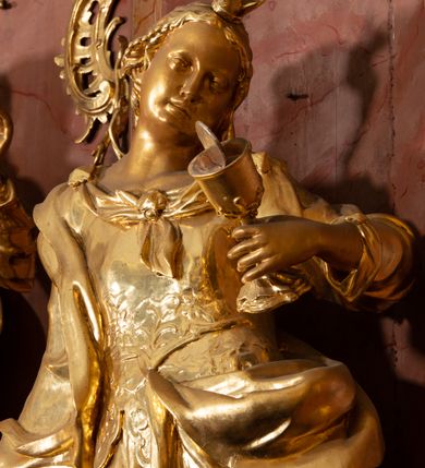 Zdjęcie nr 30: Nastawa ołtarzowa architektoniczna, jednokondygnacyjna ze zwieńczeniem, pięcioosiowa, ustawiona na wysokim cokole. Ołtarz wolnostojący, drewniany, sarkofagowy, wklęsło-wypukły, o zdwojonym froncie; mensa drewniana z marmurowym portatylem. Od frontu rocaille'owy kartusz ujęty fantazyjnymi płycinami oraz liśćmi. Na ołtarzu tabernakulum w formie sześcianu o wklęsłych bokach i wklęsło-wypukłym froncie; ujęte w narożach dwiema parami pilastrów o trzonach przechodzących w spływy wolutowe zdobione rocaille'm i różami. Pilastry podtrzymują falujący gzyms oraz fragmenty falistego, przerwanego, odwróconego przyczółka, przechodzącego w cokół, stanowiący tron wystawienia, na którym stoi krucyfiks. Na ściance frontowej obrotowa nisza, o wykroju stojącego prostokąta zamkniętego łukiem półkolistym, w opasce, zwieńczona kampanulami. W jej polu drzwiczki w formie stojącego prostokąta zamkniętego łukiem półkolistym w profilowanej ramie, z odlewanym przedstawieniem baranka leżącego na księdze z pieczęciami. Tabernakulum ujęte spływami przechodzącymi na krańcach we fragmenty wolutowego gzymsu, na których siedzą figury adorujących aniołów. Ponad tabernakulum fantazyjna ścianka tworząca tło dla tronu wystawienia z prześwitem otoczonym obłokami, z glorią promienistą, flankowana rocaille'owymi uszami. Cokół nastawy z wyodrębnionymi dwoma prostopadłościanami i dwoma konsolami, na dwóch skrajnych osiach bramki zamknięte wolutowymi szczytami tworzącymi cokoły dla figur, z przejściami w formie stojącego prostokąta zamkniętego łukiem odcinkowym nadwieszonym, z drzwiczkami. Cokół i drzwiczki dekorowane rocaille'm. Lico nastawy wklęsłe, osie wydzielone trzema parami pilastrów, na których tle stoją dwie pary kolumn o kapitelach kompozytowych. Podpory dźwigają pełne belkowanie, wyłamane na ich osi, przerwane nad polem środkowym, o gzymsie przechodzącym miejscami w rocaille'owe kartusze. Na osi środkowej wnęka z obrazem Matki Boskiej Wspomożenia Wiernych, w formie stojącego prostokąta zamkniętego łukiem odcinkowym nadwieszonym z uskokiem, ujęta profilowaną ramą, dekorowaną ponad łukiem płomienistym rocaille'm. Na pozostałych czterech osiach figury św. Barbary, św. Apolonii, św. Piotra i św. Pawła ustawione na rokokowych wspornikach. Trzony pilastrów i pola osi bocznych wypełnione rocaille'm, fryz dekorowany zwisami kwiatowymi. Zwieńczenie w formie zbliżonej do trapezu, zamknięte dwoma fragmentami gzymsu przechodzącego w woluty, flankowane wolutami spływającymi na belkowanie wewnętrznej pary kolumn, na których siedzą adorujące figury aniołów. W polu rzeźbiarska grupa Trójcy Świętej, pośród obłoków, w glorii promienistej, zwieńczona gołębicą Ducha Świętego w otoku z obłoków i uskrzydlonych główek anielskich, w glorii promienistej, flankowana przez dwa putta siedzące na gzymsie. Zwieńczenie po bokach obwiedzione rocaille'm, ujęte liśćmi palmowymi. Na belkowaniu, na osiach zewnętrznej pary kolumn wolutowe cokoły z rokokowymi wazonami. Struktura polichromowana na sposób imitujący marmoryzację w kolorze różowym i różowo-pomarańczowym, kolumny, profile i ornamentyka złocone, obłoki srebrzone. 

