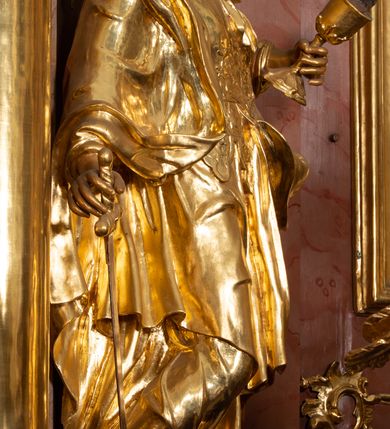 Zdjęcie nr 33: Nastawa ołtarzowa architektoniczna, jednokondygnacyjna ze zwieńczeniem, pięcioosiowa, ustawiona na wysokim cokole. Ołtarz wolnostojący, drewniany, sarkofagowy, wklęsło-wypukły, o zdwojonym froncie; mensa drewniana z marmurowym portatylem. Od frontu rocaille&#039;owy kartusz ujęty fantazyjnymi płycinami oraz liśćmi. Na ołtarzu tabernakulum w formie sześcianu o wklęsłych bokach i wklęsło-wypukłym froncie; ujęte w narożach dwiema parami pilastrów o trzonach przechodzących w spływy wolutowe zdobione rocaille&#039;m i różami. Pilastry podtrzymują falujący gzyms oraz fragmenty falistego, przerwanego, odwróconego przyczółka, przechodzącego w cokół, stanowiący tron wystawienia, na którym stoi krucyfiks. Na ściance frontowej obrotowa nisza, o wykroju stojącego prostokąta zamkniętego łukiem półkolistym, w opasce, zwieńczona kampanulami. W jej polu drzwiczki w formie stojącego prostokąta zamkniętego łukiem półkolistym w profilowanej ramie, z odlewanym przedstawieniem baranka leżącego na księdze z pieczęciami. Tabernakulum ujęte spływami przechodzącymi na krańcach we fragmenty wolutowego gzymsu, na których siedzą figury adorujących aniołów. Ponad tabernakulum fantazyjna ścianka tworząca tło dla tronu wystawienia z prześwitem otoczonym obłokami, z glorią promienistą, flankowana rocaille&#039;owymi uszami. Cokół nastawy z wyodrębnionymi dwoma prostopadłościanami i dwoma konsolami, na dwóch skrajnych osiach bramki zamknięte wolutowymi szczytami tworzącymi cokoły dla figur, z przejściami w formie stojącego prostokąta zamkniętego łukiem odcinkowym nadwieszonym, z drzwiczkami. Cokół i drzwiczki dekorowane rocaille&#039;m. Lico nastawy wklęsłe, osie wydzielone trzema parami pilastrów, na których tle stoją dwie pary kolumn o kapitelach kompozytowych. Podpory dźwigają pełne belkowanie, wyłamane na ich osi, przerwane nad polem środkowym, o gzymsie przechodzącym miejscami w rocaille&#039;owe kartusze. Na osi środkowej wnęka z obrazem Matki Boskiej Wspomożenia Wiernych, w formie stojącego prostokąta zamkniętego łukiem odcinkowym nadwieszonym z uskokiem, ujęta profilowaną ramą, dekorowaną ponad łukiem płomienistym rocaille&#039;m. Na pozostałych czterech osiach figury św. Barbary, św. Apolonii, św. Piotra i św. Pawła ustawione na rokokowych wspornikach. Trzony pilastrów i pola osi bocznych wypełnione rocaille&#039;m, fryz dekorowany zwisami kwiatowymi. Zwieńczenie w formie zbliżonej do trapezu, zamknięte dwoma fragmentami gzymsu przechodzącego w woluty, flankowane wolutami spływającymi na belkowanie wewnętrznej pary kolumn, na których siedzą adorujące figury aniołów. W polu rzeźbiarska grupa Trójcy Świętej, pośród obłoków, w glorii promienistej, zwieńczona gołębicą Ducha Świętego w otoku z obłoków i uskrzydlonych główek anielskich, w glorii promienistej, flankowana przez dwa putta siedzące na gzymsie. Zwieńczenie po bokach obwiedzione rocaille&#039;m, ujęte liśćmi palmowymi. Na belkowaniu, na osiach zewnętrznej pary kolumn wolutowe cokoły z rokokowymi wazonami. Struktura polichromowana na sposób imitujący marmoryzację w kolorze różowym i różowo-pomarańczowym, kolumny, profile i ornamentyka złocone, obłoki srebrzone. 

