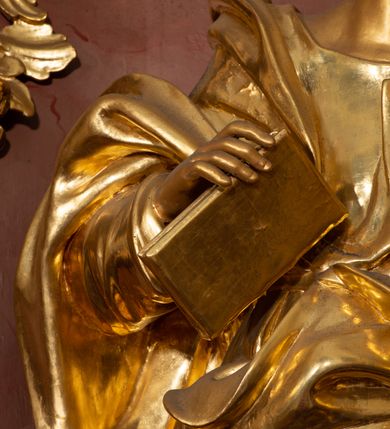 Zdjęcie nr 36: Nastawa ołtarzowa architektoniczna, jednokondygnacyjna ze zwieńczeniem, pięcioosiowa, ustawiona na wysokim cokole. Ołtarz wolnostojący, drewniany, sarkofagowy, wklęsło-wypukły, o zdwojonym froncie; mensa drewniana z marmurowym portatylem. Od frontu rocaille&#039;owy kartusz ujęty fantazyjnymi płycinami oraz liśćmi. Na ołtarzu tabernakulum w formie sześcianu o wklęsłych bokach i wklęsło-wypukłym froncie; ujęte w narożach dwiema parami pilastrów o trzonach przechodzących w spływy wolutowe zdobione rocaille&#039;m i różami. Pilastry podtrzymują falujący gzyms oraz fragmenty falistego, przerwanego, odwróconego przyczółka, przechodzącego w cokół, stanowiący tron wystawienia, na którym stoi krucyfiks. Na ściance frontowej obrotowa nisza, o wykroju stojącego prostokąta zamkniętego łukiem półkolistym, w opasce, zwieńczona kampanulami. W jej polu drzwiczki w formie stojącego prostokąta zamkniętego łukiem półkolistym w profilowanej ramie, z odlewanym przedstawieniem baranka leżącego na księdze z pieczęciami. Tabernakulum ujęte spływami przechodzącymi na krańcach we fragmenty wolutowego gzymsu, na których siedzą figury adorujących aniołów. Ponad tabernakulum fantazyjna ścianka tworząca tło dla tronu wystawienia z prześwitem otoczonym obłokami, z glorią promienistą, flankowana rocaille&#039;owymi uszami. Cokół nastawy z wyodrębnionymi dwoma prostopadłościanami i dwoma konsolami, na dwóch skrajnych osiach bramki zamknięte wolutowymi szczytami tworzącymi cokoły dla figur, z przejściami w formie stojącego prostokąta zamkniętego łukiem odcinkowym nadwieszonym, z drzwiczkami. Cokół i drzwiczki dekorowane rocaille&#039;m. Lico nastawy wklęsłe, osie wydzielone trzema parami pilastrów, na których tle stoją dwie pary kolumn o kapitelach kompozytowych. Podpory dźwigają pełne belkowanie, wyłamane na ich osi, przerwane nad polem środkowym, o gzymsie przechodzącym miejscami w rocaille&#039;owe kartusze. Na osi środkowej wnęka z obrazem Matki Boskiej Wspomożenia Wiernych, w formie stojącego prostokąta zamkniętego łukiem odcinkowym nadwieszonym z uskokiem, ujęta profilowaną ramą, dekorowaną ponad łukiem płomienistym rocaille&#039;m. Na pozostałych czterech osiach figury św. Barbary, św. Apolonii, św. Piotra i św. Pawła ustawione na rokokowych wspornikach. Trzony pilastrów i pola osi bocznych wypełnione rocaille&#039;m, fryz dekorowany zwisami kwiatowymi. Zwieńczenie w formie zbliżonej do trapezu, zamknięte dwoma fragmentami gzymsu przechodzącego w woluty, flankowane wolutami spływającymi na belkowanie wewnętrznej pary kolumn, na których siedzą adorujące figury aniołów. W polu rzeźbiarska grupa Trójcy Świętej, pośród obłoków, w glorii promienistej, zwieńczona gołębicą Ducha Świętego w otoku z obłoków i uskrzydlonych główek anielskich, w glorii promienistej, flankowana przez dwa putta siedzące na gzymsie. Zwieńczenie po bokach obwiedzione rocaille&#039;m, ujęte liśćmi palmowymi. Na belkowaniu, na osiach zewnętrznej pary kolumn wolutowe cokoły z rokokowymi wazonami. Struktura polichromowana na sposób imitujący marmoryzację w kolorze różowym i różowo-pomarańczowym, kolumny, profile i ornamentyka złocone, obłoki srebrzone. 

