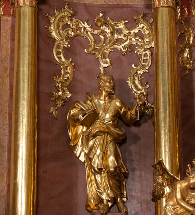 Zdjęcie nr 39: Nastawa ołtarzowa architektoniczna, jednokondygnacyjna ze zwieńczeniem, pięcioosiowa, ustawiona na wysokim cokole. Ołtarz wolnostojący, drewniany, sarkofagowy, wklęsło-wypukły, o zdwojonym froncie; mensa drewniana z marmurowym portatylem. Od frontu rocaille'owy kartusz ujęty fantazyjnymi płycinami oraz liśćmi. Na ołtarzu tabernakulum w formie sześcianu o wklęsłych bokach i wklęsło-wypukłym froncie; ujęte w narożach dwiema parami pilastrów o trzonach przechodzących w spływy wolutowe zdobione rocaille'm i różami. Pilastry podtrzymują falujący gzyms oraz fragmenty falistego, przerwanego, odwróconego przyczółka, przechodzącego w cokół, stanowiący tron wystawienia, na którym stoi krucyfiks. Na ściance frontowej obrotowa nisza, o wykroju stojącego prostokąta zamkniętego łukiem półkolistym, w opasce, zwieńczona kampanulami. W jej polu drzwiczki w formie stojącego prostokąta zamkniętego łukiem półkolistym w profilowanej ramie, z odlewanym przedstawieniem baranka leżącego na księdze z pieczęciami. Tabernakulum ujęte spływami przechodzącymi na krańcach we fragmenty wolutowego gzymsu, na których siedzą figury adorujących aniołów. Ponad tabernakulum fantazyjna ścianka tworząca tło dla tronu wystawienia z prześwitem otoczonym obłokami, z glorią promienistą, flankowana rocaille'owymi uszami. Cokół nastawy z wyodrębnionymi dwoma prostopadłościanami i dwoma konsolami, na dwóch skrajnych osiach bramki zamknięte wolutowymi szczytami tworzącymi cokoły dla figur, z przejściami w formie stojącego prostokąta zamkniętego łukiem odcinkowym nadwieszonym, z drzwiczkami. Cokół i drzwiczki dekorowane rocaille'm. Lico nastawy wklęsłe, osie wydzielone trzema parami pilastrów, na których tle stoją dwie pary kolumn o kapitelach kompozytowych. Podpory dźwigają pełne belkowanie, wyłamane na ich osi, przerwane nad polem środkowym, o gzymsie przechodzącym miejscami w rocaille'owe kartusze. Na osi środkowej wnęka z obrazem Matki Boskiej Wspomożenia Wiernych, w formie stojącego prostokąta zamkniętego łukiem odcinkowym nadwieszonym z uskokiem, ujęta profilowaną ramą, dekorowaną ponad łukiem płomienistym rocaille'm. Na pozostałych czterech osiach figury św. Barbary, św. Apolonii, św. Piotra i św. Pawła ustawione na rokokowych wspornikach. Trzony pilastrów i pola osi bocznych wypełnione rocaille'm, fryz dekorowany zwisami kwiatowymi. Zwieńczenie w formie zbliżonej do trapezu, zamknięte dwoma fragmentami gzymsu przechodzącego w woluty, flankowane wolutami spływającymi na belkowanie wewnętrznej pary kolumn, na których siedzą adorujące figury aniołów. W polu rzeźbiarska grupa Trójcy Świętej, pośród obłoków, w glorii promienistej, zwieńczona gołębicą Ducha Świętego w otoku z obłoków i uskrzydlonych główek anielskich, w glorii promienistej, flankowana przez dwa putta siedzące na gzymsie. Zwieńczenie po bokach obwiedzione rocaille'm, ujęte liśćmi palmowymi. Na belkowaniu, na osiach zewnętrznej pary kolumn wolutowe cokoły z rokokowymi wazonami. Struktura polichromowana na sposób imitujący marmoryzację w kolorze różowym i różowo-pomarańczowym, kolumny, profile i ornamentyka złocone, obłoki srebrzone. 

