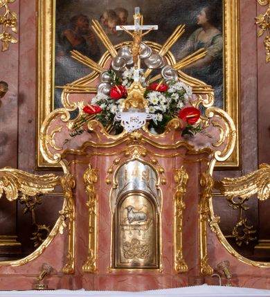 Zdjęcie nr 54: Nastawa ołtarzowa architektoniczna, jednokondygnacyjna ze zwieńczeniem, pięcioosiowa, ustawiona na wysokim cokole. Ołtarz wolnostojący, drewniany, sarkofagowy, wklęsło-wypukły, o zdwojonym froncie; mensa drewniana z marmurowym portatylem. Od frontu rocaille&#039;owy kartusz ujęty fantazyjnymi płycinami oraz liśćmi. Na ołtarzu tabernakulum w formie sześcianu o wklęsłych bokach i wklęsło-wypukłym froncie; ujęte w narożach dwiema parami pilastrów o trzonach przechodzących w spływy wolutowe zdobione rocaille&#039;m i różami. Pilastry podtrzymują falujący gzyms oraz fragmenty falistego, przerwanego, odwróconego przyczółka, przechodzącego w cokół, stanowiący tron wystawienia, na którym stoi krucyfiks. Na ściance frontowej obrotowa nisza, o wykroju stojącego prostokąta zamkniętego łukiem półkolistym, w opasce, zwieńczona kampanulami. W jej polu drzwiczki w formie stojącego prostokąta zamkniętego łukiem półkolistym w profilowanej ramie, z odlewanym przedstawieniem baranka leżącego na księdze z pieczęciami. Tabernakulum ujęte spływami przechodzącymi na krańcach we fragmenty wolutowego gzymsu, na których siedzą figury adorujących aniołów. Ponad tabernakulum fantazyjna ścianka tworząca tło dla tronu wystawienia z prześwitem otoczonym obłokami, z glorią promienistą, flankowana rocaille&#039;owymi uszami. Cokół nastawy z wyodrębnionymi dwoma prostopadłościanami i dwoma konsolami, na dwóch skrajnych osiach bramki zamknięte wolutowymi szczytami tworzącymi cokoły dla figur, z przejściami w formie stojącego prostokąta zamkniętego łukiem odcinkowym nadwieszonym, z drzwiczkami. Cokół i drzwiczki dekorowane rocaille&#039;m. Lico nastawy wklęsłe, osie wydzielone trzema parami pilastrów, na których tle stoją dwie pary kolumn o kapitelach kompozytowych. Podpory dźwigają pełne belkowanie, wyłamane na ich osi, przerwane nad polem środkowym, o gzymsie przechodzącym miejscami w rocaille&#039;owe kartusze. Na osi środkowej wnęka z obrazem Matki Boskiej Wspomożenia Wiernych, w formie stojącego prostokąta zamkniętego łukiem odcinkowym nadwieszonym z uskokiem, ujęta profilowaną ramą, dekorowaną ponad łukiem płomienistym rocaille&#039;m. Na pozostałych czterech osiach figury św. Barbary, św. Apolonii, św. Piotra i św. Pawła ustawione na rokokowych wspornikach. Trzony pilastrów i pola osi bocznych wypełnione rocaille&#039;m, fryz dekorowany zwisami kwiatowymi. Zwieńczenie w formie zbliżonej do trapezu, zamknięte dwoma fragmentami gzymsu przechodzącego w woluty, flankowane wolutami spływającymi na belkowanie wewnętrznej pary kolumn, na których siedzą adorujące figury aniołów. W polu rzeźbiarska grupa Trójcy Świętej, pośród obłoków, w glorii promienistej, zwieńczona gołębicą Ducha Świętego w otoku z obłoków i uskrzydlonych główek anielskich, w glorii promienistej, flankowana przez dwa putta siedzące na gzymsie. Zwieńczenie po bokach obwiedzione rocaille&#039;m, ujęte liśćmi palmowymi. Na belkowaniu, na osiach zewnętrznej pary kolumn wolutowe cokoły z rokokowymi wazonami. Struktura polichromowana na sposób imitujący marmoryzację w kolorze różowym i różowo-pomarańczowym, kolumny, profile i ornamentyka złocone, obłoki srebrzone. 

