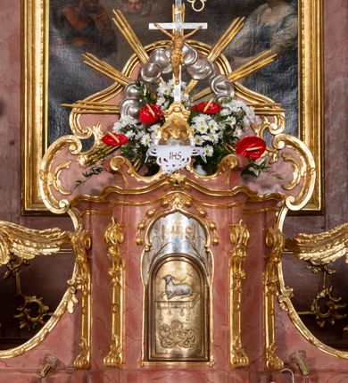 Zdjęcie nr 55: Nastawa ołtarzowa architektoniczna, jednokondygnacyjna ze zwieńczeniem, pięcioosiowa, ustawiona na wysokim cokole. Ołtarz wolnostojący, drewniany, sarkofagowy, wklęsło-wypukły, o zdwojonym froncie; mensa drewniana z marmurowym portatylem. Od frontu rocaille'owy kartusz ujęty fantazyjnymi płycinami oraz liśćmi. Na ołtarzu tabernakulum w formie sześcianu o wklęsłych bokach i wklęsło-wypukłym froncie; ujęte w narożach dwiema parami pilastrów o trzonach przechodzących w spływy wolutowe zdobione rocaille'm i różami. Pilastry podtrzymują falujący gzyms oraz fragmenty falistego, przerwanego, odwróconego przyczółka, przechodzącego w cokół, stanowiący tron wystawienia, na którym stoi krucyfiks. Na ściance frontowej obrotowa nisza, o wykroju stojącego prostokąta zamkniętego łukiem półkolistym, w opasce, zwieńczona kampanulami. W jej polu drzwiczki w formie stojącego prostokąta zamkniętego łukiem półkolistym w profilowanej ramie, z odlewanym przedstawieniem baranka leżącego na księdze z pieczęciami. Tabernakulum ujęte spływami przechodzącymi na krańcach we fragmenty wolutowego gzymsu, na których siedzą figury adorujących aniołów. Ponad tabernakulum fantazyjna ścianka tworząca tło dla tronu wystawienia z prześwitem otoczonym obłokami, z glorią promienistą, flankowana rocaille'owymi uszami. Cokół nastawy z wyodrębnionymi dwoma prostopadłościanami i dwoma konsolami, na dwóch skrajnych osiach bramki zamknięte wolutowymi szczytami tworzącymi cokoły dla figur, z przejściami w formie stojącego prostokąta zamkniętego łukiem odcinkowym nadwieszonym, z drzwiczkami. Cokół i drzwiczki dekorowane rocaille'm. Lico nastawy wklęsłe, osie wydzielone trzema parami pilastrów, na których tle stoją dwie pary kolumn o kapitelach kompozytowych. Podpory dźwigają pełne belkowanie, wyłamane na ich osi, przerwane nad polem środkowym, o gzymsie przechodzącym miejscami w rocaille'owe kartusze. Na osi środkowej wnęka z obrazem Matki Boskiej Wspomożenia Wiernych, w formie stojącego prostokąta zamkniętego łukiem odcinkowym nadwieszonym z uskokiem, ujęta profilowaną ramą, dekorowaną ponad łukiem płomienistym rocaille'm. Na pozostałych czterech osiach figury św. Barbary, św. Apolonii, św. Piotra i św. Pawła ustawione na rokokowych wspornikach. Trzony pilastrów i pola osi bocznych wypełnione rocaille'm, fryz dekorowany zwisami kwiatowymi. Zwieńczenie w formie zbliżonej do trapezu, zamknięte dwoma fragmentami gzymsu przechodzącego w woluty, flankowane wolutami spływającymi na belkowanie wewnętrznej pary kolumn, na których siedzą adorujące figury aniołów. W polu rzeźbiarska grupa Trójcy Świętej, pośród obłoków, w glorii promienistej, zwieńczona gołębicą Ducha Świętego w otoku z obłoków i uskrzydlonych główek anielskich, w glorii promienistej, flankowana przez dwa putta siedzące na gzymsie. Zwieńczenie po bokach obwiedzione rocaille'm, ujęte liśćmi palmowymi. Na belkowaniu, na osiach zewnętrznej pary kolumn wolutowe cokoły z rokokowymi wazonami. Struktura polichromowana na sposób imitujący marmoryzację w kolorze różowym i różowo-pomarańczowym, kolumny, profile i ornamentyka złocone, obłoki srebrzone. 

