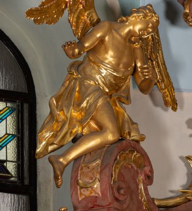 Zdjęcie nr 52: Nastawa ołtarzowa architektoniczna, jednokondygnacyjna ze zwieńczeniem, pięcioosiowa, ustawiona na wysokim cokole. Ołtarz wolnostojący, drewniany, sarkofagowy, wklęsło-wypukły, o zdwojonym froncie; mensa drewniana z marmurowym portatylem. Od frontu rocaille&#039;owy kartusz ujęty fantazyjnymi płycinami oraz liśćmi. Na ołtarzu tabernakulum w formie sześcianu o wklęsłych bokach i wklęsło-wypukłym froncie; ujęte w narożach dwiema parami pilastrów o trzonach przechodzących w spływy wolutowe zdobione rocaille&#039;m i różami. Pilastry podtrzymują falujący gzyms oraz fragmenty falistego, przerwanego, odwróconego przyczółka, przechodzącego w cokół, stanowiący tron wystawienia, na którym stoi krucyfiks. Na ściance frontowej obrotowa nisza, o wykroju stojącego prostokąta zamkniętego łukiem półkolistym, w opasce, zwieńczona kampanulami. W jej polu drzwiczki w formie stojącego prostokąta zamkniętego łukiem półkolistym w profilowanej ramie, z odlewanym przedstawieniem baranka leżącego na księdze z pieczęciami. Tabernakulum ujęte spływami przechodzącymi na krańcach we fragmenty wolutowego gzymsu, na których siedzą figury adorujących aniołów. Ponad tabernakulum fantazyjna ścianka tworząca tło dla tronu wystawienia z prześwitem otoczonym obłokami, z glorią promienistą, flankowana rocaille&#039;owymi uszami. Cokół nastawy z wyodrębnionymi dwoma prostopadłościanami i dwoma konsolami, na dwóch skrajnych osiach bramki zamknięte wolutowymi szczytami tworzącymi cokoły dla figur, z przejściami w formie stojącego prostokąta zamkniętego łukiem odcinkowym nadwieszonym, z drzwiczkami. Cokół i drzwiczki dekorowane rocaille&#039;m. Lico nastawy wklęsłe, osie wydzielone trzema parami pilastrów, na których tle stoją dwie pary kolumn o kapitelach kompozytowych. Podpory dźwigają pełne belkowanie, wyłamane na ich osi, przerwane nad polem środkowym, o gzymsie przechodzącym miejscami w rocaille&#039;owe kartusze. Na osi środkowej wnęka z obrazem Matki Boskiej Wspomożenia Wiernych, w formie stojącego prostokąta zamkniętego łukiem odcinkowym nadwieszonym z uskokiem, ujęta profilowaną ramą, dekorowaną ponad łukiem płomienistym rocaille&#039;m. Na pozostałych czterech osiach figury św. Barbary, św. Apolonii, św. Piotra i św. Pawła ustawione na rokokowych wspornikach. Trzony pilastrów i pola osi bocznych wypełnione rocaille&#039;m, fryz dekorowany zwisami kwiatowymi. Zwieńczenie w formie zbliżonej do trapezu, zamknięte dwoma fragmentami gzymsu przechodzącego w woluty, flankowane wolutami spływającymi na belkowanie wewnętrznej pary kolumn, na których siedzą adorujące figury aniołów. W polu rzeźbiarska grupa Trójcy Świętej, pośród obłoków, w glorii promienistej, zwieńczona gołębicą Ducha Świętego w otoku z obłoków i uskrzydlonych główek anielskich, w glorii promienistej, flankowana przez dwa putta siedzące na gzymsie. Zwieńczenie po bokach obwiedzione rocaille&#039;m, ujęte liśćmi palmowymi. Na belkowaniu, na osiach zewnętrznej pary kolumn wolutowe cokoły z rokokowymi wazonami. Struktura polichromowana na sposób imitujący marmoryzację w kolorze różowym i różowo-pomarańczowym, kolumny, profile i ornamentyka złocone, obłoki srebrzone. 

