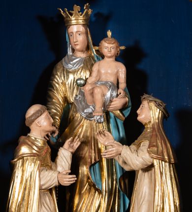 Zdjęcie nr 1: Grupa rzeźbiarska przedstawiająca stojącą Matkę Boską z Dzieciątkiem, przed którą klęczą święci. W centrum figura Marii, ustawiona na cokole, ukazana frontalnie, w całej postaci, z głową skierowaną w prawo. Lewą ręką podtrzymuje Dzieciątko, prawą wyciąga w stronę św. Dominika. Twarz ma pociągłą, o migdałowatych oczach, prostym nosie i wąskich ustach, okoloną brązowymi włosami. Ubrana jest w złoconą tunikę i złocony płaszcz o niebieskiej podszewce, zarzucony na głowę i opasujący postać. Na głowie ma otwartą koronę. Dzieciątko w pozycji siedzącej, zwrócone w trzech czwartych w prawo, z głową ukazaną frontalnie, w prawej ręce trzyma jabłko królewskie, lewą wyciąga w stronę św. Katarzyny. Twarz ma kwadratową, o wąskich oczach, krótkim nosie i małych ustach, okoloną jasnobrązowymi, krótkimi włosami. Z jego głowy odchodzą trzy wiązki promieni. W partii bioder przewiązane jest srebrzoną pieluszką. Po prawej stronie Marii klęczy św. Dominik, ukazany z profilu, z uniesioną głową i wyciągniętymi przed siebie rękami. Twarz ma szczupłą, o migdałowatych oczach, prostym nosie i małych ustach, okoloną krótkimi włosami, z tonsurą na głowie. Ubrany jest w biały habit, ze złoconą pelerynką, szkaplerzem oraz płaszczem. Po lewej stronie Marii klęczy św. Katarzyna, ukazana z profilu, z uniesioną głową i wyciągniętymi przed siebie rękami. Twarz ma szczupłą, o migdałowatych oczach, prostym nosie i małych ustach. Ubrana jest w biały habit, ze złoconą pelerynką, szkaplerzem, welonem i płaszczem. Na głowie ma koronę cierniową. Polichromia ciał postaci naturalistyczna. 

