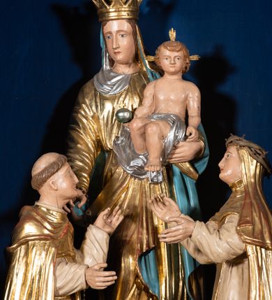 Zdjęcie nr 2: Grupa rzeźbiarska przedstawiająca stojącą Matkę Boską z Dzieciątkiem, przed którą klęczą święci. W centrum figura Marii, ustawiona na cokole, ukazana frontalnie, w całej postaci, z głową skierowaną w prawo. Lewą ręką podtrzymuje Dzieciątko, prawą wyciąga w stronę św. Dominika. Twarz ma pociągłą, o migdałowatych oczach, prostym nosie i wąskich ustach, okoloną brązowymi włosami. Ubrana jest w złoconą tunikę i złocony płaszcz o niebieskiej podszewce, zarzucony na głowę i opasujący postać. Na głowie ma otwartą koronę. Dzieciątko w pozycji siedzącej, zwrócone w trzech czwartych w prawo, z głową ukazaną frontalnie, w prawej ręce trzyma jabłko królewskie, lewą wyciąga w stronę św. Katarzyny. Twarz ma kwadratową, o wąskich oczach, krótkim nosie i małych ustach, okoloną jasnobrązowymi, krótkimi włosami. Z jego głowy odchodzą trzy wiązki promieni. W partii bioder przewiązane jest srebrzoną pieluszką. Po prawej stronie Marii klęczy św. Dominik, ukazany z profilu, z uniesioną głową i wyciągniętymi przed siebie rękami. Twarz ma szczupłą, o migdałowatych oczach, prostym nosie i małych ustach, okoloną krótkimi włosami, z tonsurą na głowie. Ubrany jest w biały habit, ze złoconą pelerynką, szkaplerzem oraz płaszczem. Po lewej stronie Marii klęczy św. Katarzyna, ukazana z profilu, z uniesioną głową i wyciągniętymi przed siebie rękami. Twarz ma szczupłą, o migdałowatych oczach, prostym nosie i małych ustach. Ubrana jest w biały habit, ze złoconą pelerynką, szkaplerzem, welonem i płaszczem. Na głowie ma koronę cierniową. Polichromia ciał postaci naturalistyczna. 

