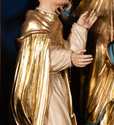 Zdjęcie nr 3: Grupa rzeźbiarska przedstawiająca stojącą Matkę Boską z Dzieciątkiem, przed którą klęczą święci. W centrum figura Marii, ustawiona na cokole, ukazana frontalnie, w całej postaci, z głową skierowaną w prawo. Lewą ręką podtrzymuje Dzieciątko, prawą wyciąga w stronę św. Dominika. Twarz ma pociągłą, o migdałowatych oczach, prostym nosie i wąskich ustach, okoloną brązowymi włosami. Ubrana jest w złoconą tunikę i złocony płaszcz o niebieskiej podszewce, zarzucony na głowę i opasujący postać. Na głowie ma otwartą koronę. Dzieciątko w pozycji siedzącej, zwrócone w trzech czwartych w prawo, z głową ukazaną frontalnie, w prawej ręce trzyma jabłko królewskie, lewą wyciąga w stronę św. Katarzyny. Twarz ma kwadratową, o wąskich oczach, krótkim nosie i małych ustach, okoloną jasnobrązowymi, krótkimi włosami. Z jego głowy odchodzą trzy wiązki promieni. W partii bioder przewiązane jest srebrzoną pieluszką. Po prawej stronie Marii klęczy św. Dominik, ukazany z profilu, z uniesioną głową i wyciągniętymi przed siebie rękami. Twarz ma szczupłą, o migdałowatych oczach, prostym nosie i małych ustach, okoloną krótkimi włosami, z tonsurą na głowie. Ubrany jest w biały habit, ze złoconą pelerynką, szkaplerzem oraz płaszczem. Po lewej stronie Marii klęczy św. Katarzyna, ukazana z profilu, z uniesioną głową i wyciągniętymi przed siebie rękami. Twarz ma szczupłą, o migdałowatych oczach, prostym nosie i małych ustach. Ubrana jest w biały habit, ze złoconą pelerynką, szkaplerzem, welonem i płaszczem. Na głowie ma koronę cierniową. Polichromia ciał postaci naturalistyczna. 

