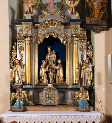 Zdjęcie nr 5: Grupa rzeźbiarska przedstawiająca stojącą Matkę Boską z Dzieciątkiem, przed którą klęczą święci. W centrum figura Marii, ustawiona na cokole, ukazana frontalnie, w całej postaci, z głową skierowaną w prawo. Lewą ręką podtrzymuje Dzieciątko, prawą wyciąga w stronę św. Dominika. Twarz ma pociągłą, o migdałowatych oczach, prostym nosie i wąskich ustach, okoloną brązowymi włosami. Ubrana jest w złoconą tunikę i złocony płaszcz o niebieskiej podszewce, zarzucony na głowę i opasujący postać. Na głowie ma otwartą koronę. Dzieciątko w pozycji siedzącej, zwrócone w trzech czwartych w prawo, z głową ukazaną frontalnie, w prawej ręce trzyma jabłko królewskie, lewą wyciąga w stronę św. Katarzyny. Twarz ma kwadratową, o wąskich oczach, krótkim nosie i małych ustach, okoloną jasnobrązowymi, krótkimi włosami. Z jego głowy odchodzą trzy wiązki promieni. W partii bioder przewiązane jest srebrzoną pieluszką. Po prawej stronie Marii klęczy św. Dominik, ukazany z profilu, z uniesioną głową i wyciągniętymi przed siebie rękami. Twarz ma szczupłą, o migdałowatych oczach, prostym nosie i małych ustach, okoloną krótkimi włosami, z tonsurą na głowie. Ubrany jest w biały habit, ze złoconą pelerynką, szkaplerzem oraz płaszczem. Po lewej stronie Marii klęczy św. Katarzyna, ukazana z profilu, z uniesioną głową i wyciągniętymi przed siebie rękami. Twarz ma szczupłą, o migdałowatych oczach, prostym nosie i małych ustach. Ubrana jest w biały habit, ze złoconą pelerynką, szkaplerzem, welonem i płaszczem. Na głowie ma koronę cierniową. Polichromia ciał postaci naturalistyczna. 

