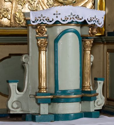 Zdjęcie nr 10: Nastawa architektoniczna, jednokondygnacyjna, jednoosiowa, ustawiona na cokole z wyodrębnionymi prostopadłościanami. Ołtarz drewniany, sarkofagowy, na cokole, z drewnianą mensą i kamiennym portatylem. Cokół nastawy dekorowany płycinami z akantem. Retabulum ujęte parą kolumn o skręconych trzonach i korynckich kapitelach, dźwigających pełne belkowanie, nad polem środkowym wyłamane na kształt łuku odcinkowego. Na belkowaniu fragmenty falistego, przerwanego przyczółka z siedzącymi puttami. Całe pole środkowe wypełnione wnęką w formie stojącego prostokąta zamkniętego łukiem półkolistym nadwieszonym, ujętą profilowaną ramą, z obrazem Matki Boskiej Wspomożenia Wiernych. Nastawa flankowana uszami utworzonymi z akantu i kratki regencyjnej. W miejscu zwieńczenia figura siedzącego na obłoku Chrystusa trzymającego krzyż, w glorii promienistej. Struktura polichromowana w kolorze beżowym, z jasnoniebieskimi i niebieskimi detalami, złoconymi kolumnami, profilami i ornamentyką oraz srebrzonymi detalami. 
