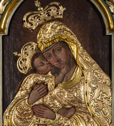 Zdjęcie nr 1: Obraz w formie stojącego prostokąta zamkniętego łukiem półkolistym nadwieszonym. Postacie przesłonięte są drewnianą sukienką w partiach szat, odsłaniającą twarze, dłonie i nogi. W centrum Matka Boska ukazana do kolan, zwrócona w trzech czwartych w prawo, obiema rękami obejmuje Dzieciątko, przytulając swoją twarz do jego policzka. Twarz ma trójkątną, o migdałowatych oczach, łukowato wygiętych brwiach, prostym, wąskim nosie i małych ustach. Ubrana jest w suknię i płaszcz nakrywający głowę i opadający na kolana, przesłonięte sukienką o płaskorzeźbionym roślinnym wzorze, z płaszczem o dekoracyjnej lamówce imitującej kameryzację. Dzieciątko ukazane z profilu, lewą nogą stoi na kolanach Matki, prawą unosi, przytula twarz do Marii i obejmuje ją prawą ręką. Twarz ma okrągłą, o dużych, skierowanych w górę oczach, małym, zadartym nosie i pełnych ustach, okoloną krótkimi, brązowymi włosami. Jego ciało przesłonięte jest drewnianą sukienką, w formie tuniki o płaskorzeźbionym, roślinnym wzorze, odsłaniającą nogi i rękę. Nad głowami obu postaci aplikowane metalowe korony zamknięte. Postacie ukazane na ciemnobrązowym tle, rozjaśnionym wokół postaci Marii. 



