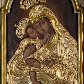 Zdjęcie nr 1: Obraz w formie stojącego prostokąta zamkniętego łukiem półkolistym nadwieszonym. Postacie przesłonięte są drewnianą sukienką w partiach szat, odsłaniającą twarze, dłonie i nogi. W centrum Matka Boska ukazana do kolan, zwrócona w trzech czwartych w prawo, obiema rękami obejmuje Dzieciątko, przytulając swoją twarz do jego policzka. Twarz ma trójkątną, o migdałowatych oczach, łukowato wygiętych brwiach, prostym, wąskim nosie i małych ustach. Ubrana jest w suknię i płaszcz nakrywający głowę i opadający na kolana, przesłonięte sukienką o płaskorzeźbionym roślinnym wzorze, z płaszczem o dekoracyjnej lamówce imitującej kameryzację. Dzieciątko ukazane z profilu, lewą nogą stoi na kolanach Matki, prawą unosi, przytula twarz do Marii i obejmuje ją prawą ręką. Twarz ma okrągłą, o dużych, skierowanych w górę oczach, małym, zadartym nosie i pełnych ustach, okoloną krótkimi, brązowymi włosami. Jego ciało przesłonięte jest drewnianą sukienką, w formie tuniki o płaskorzeźbionym, roślinnym wzorze, odsłaniającą nogi i rękę. Nad głowami obu postaci aplikowane metalowe korony zamknięte. Postacie ukazane na ciemnobrązowym tle, rozjaśnionym wokół postaci Marii. 



