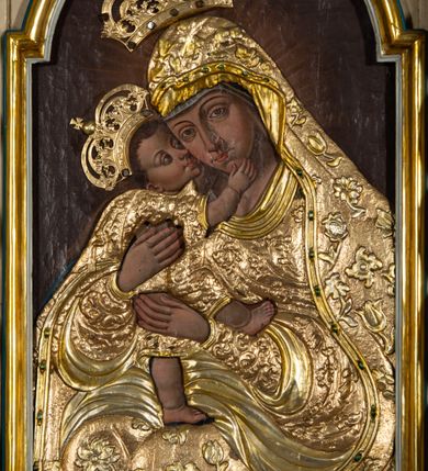 Zdjęcie nr 3: Obraz w formie stojącego prostokąta zamkniętego łukiem półkolistym nadwieszonym. Postacie przesłonięte są drewnianą sukienką w partiach szat, odsłaniającą twarze, dłonie i nogi. W centrum Matka Boska ukazana do kolan, zwrócona w trzech czwartych w prawo, obiema rękami obejmuje Dzieciątko, przytulając swoją twarz do jego policzka. Twarz ma trójkątną, o migdałowatych oczach, łukowato wygiętych brwiach, prostym, wąskim nosie i małych ustach. Ubrana jest w suknię i płaszcz nakrywający głowę i opadający na kolana, przesłonięte sukienką o płaskorzeźbionym roślinnym wzorze, z płaszczem o dekoracyjnej lamówce imitującej kameryzację. Dzieciątko ukazane z profilu, lewą nogą stoi na kolanach Matki, prawą unosi, przytula twarz do Marii i obejmuje ją prawą ręką. Twarz ma okrągłą, o dużych, skierowanych w górę oczach, małym, zadartym nosie i pełnych ustach, okoloną krótkimi, brązowymi włosami. Jego ciało przesłonięte jest drewnianą sukienką, w formie tuniki o płaskorzeźbionym, roślinnym wzorze, odsłaniającą nogi i rękę. Nad głowami obu postaci aplikowane metalowe korony zamknięte. Postacie ukazane na ciemnobrązowym tle, rozjaśnionym wokół postaci Marii. 



