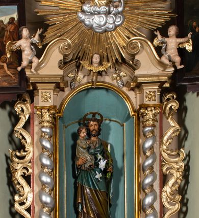 Zdjęcie nr 2: Nastawa architektoniczna, jednokondygnacyjna, jednoosiowa, ustawiona na cokole z wyodrębnionymi prostopadłościanami. Ołtarz drewniany, sarkofagowy, na cokole, z drewnianą mensą i kamiennym portatylem. Na ołtarzu ustawiony postument, ujęty spływami wolutowymi, dekorowany z przodu akantem i muszlą. Cokół nastawy dekorowany płycinami z uskrzydlonymi główkami anielskimi. Retabulum ujęte parą kolumn o skręconych trzonach i korynckich kapitelach, dźwigających niepełne belkowanie, częściowo przerwane nad polem środkowym, na którym gzyms wyłamany jest na kształt łuku odcinkowego. Na belkowaniu fragmenty falistego, przerwanego przyczółka z siedzącymi puttami. Całe pole środkowe wypełnione niszą w formie stojącego prostokąta zamkniętego łukiem półkolistym nadwieszonym, ujętą profilowaną ramą, z figurą św. Józefa z Dzieciątkiem. Nad niszą uskrzydlona główka anielska i dwie kiści winogron. Nastawa flankowana uszami utworzonymi z akantu i wstęgi regencyjnej. W miejscu zwieńczenia płaskorzeźbiona półpostać Boga Ojca pośród obłoków, w glorii promienistej. Struktura polichromowana w kolorze beżowym, z jasnoróżowymi detalami, srebrzonymi trzonami kolumn oraz złoconymi profilami i ornamentyką. 
