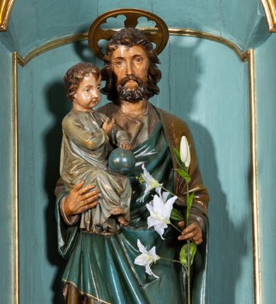 Zdjęcie nr 1: Rzeźba pełnoplastyczna, ustawiona na niskim, ośmiobocznym postumencie. Święty ukazany w całej postaci, frontalnie, w kontrapoście, prawą ręką podtrzymuje na ramieniu Dzieciątko, w lewej, ugiętej trzyma gałązkę lilii. Twarz ma szczupłą, pociągłą, o małych, migdałowatych oczach, prostym nosie i małych ustach, okoloną krótką brodą oraz kręconymi, brązowymi włosami. Nad głową nimb. Ubrany jest w lejącą szaro-brązową tunikę oraz brązowo-zielony płaszcz z niebieską podszewką, lamowany złoconą dekoracją roślinną, zarzucony na ramiona i zawinięty wokół ciała. Dzieciątko w pozycji siedzącej, zwrócone w trzech czwartych w lewo, prawą ręką wskazuje na Józefa, lewą przytrzymuje na kolanach kulę. Twarz ma pociągłą, o pełnych policzkach, dużych, migdałowatych oczach, małym nosie i ustach, okoloną krótkimi, brązowymi włosami. Ubrane jest w luźną jasnoniebieską tunikę pokrytą złoconymi gwiazdami, ze złoconą lamówką. Polichromia ciał naturalistyczna. 


