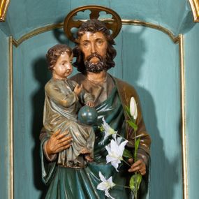 Zdjęcie nr 1: Rzeźba pełnoplastyczna, ustawiona na niskim, ośmiobocznym postumencie. Święty ukazany w całej postaci, frontalnie, w kontrapoście, prawą ręką podtrzymuje na ramieniu Dzieciątko, w lewej, ugiętej trzyma gałązkę lilii. Twarz ma szczupłą, pociągłą, o małych, migdałowatych oczach, prostym nosie i małych ustach, okoloną krótką brodą oraz kręconymi, brązowymi włosami. Nad głową nimb. Ubrany jest w lejącą szaro-brązową tunikę oraz brązowo-zielony płaszcz z niebieską podszewką, lamowany złoconą dekoracją roślinną, zarzucony na ramiona i zawinięty wokół ciała. Dzieciątko w pozycji siedzącej, zwrócone w trzech czwartych w lewo, prawą ręką wskazuje na Józefa, lewą przytrzymuje na kolanach kulę. Twarz ma pociągłą, o pełnych policzkach, dużych, migdałowatych oczach, małym nosie i ustach, okoloną krótkimi, brązowymi włosami. Ubrane jest w luźną jasnoniebieską tunikę pokrytą złoconymi gwiazdami, ze złoconą lamówką. Polichromia ciał naturalistyczna. 


