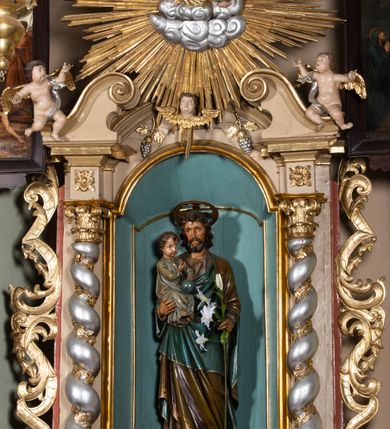 Zdjęcie nr 2: Rzeźba pełnoplastyczna, ustawiona na niskim, ośmiobocznym postumencie. Święty ukazany w całej postaci, frontalnie, w kontrapoście, prawą ręką podtrzymuje na ramieniu Dzieciątko, w lewej, ugiętej trzyma gałązkę lilii. Twarz ma szczupłą, pociągłą, o małych, migdałowatych oczach, prostym nosie i małych ustach, okoloną krótką brodą oraz kręconymi, brązowymi włosami. Nad głową nimb. Ubrany jest w lejącą szaro-brązową tunikę oraz brązowo-zielony płaszcz z niebieską podszewką, lamowany złoconą dekoracją roślinną, zarzucony na ramiona i zawinięty wokół ciała. Dzieciątko w pozycji siedzącej, zwrócone w trzech czwartych w lewo, prawą ręką wskazuje na Józefa, lewą przytrzymuje na kolanach kulę. Twarz ma pociągłą, o pełnych policzkach, dużych, migdałowatych oczach, małym nosie i ustach, okoloną krótkimi, brązowymi włosami. Ubrane jest w luźną jasnoniebieską tunikę pokrytą złoconymi gwiazdami, ze złoconą lamówką. Polichromia ciał naturalistyczna. 


