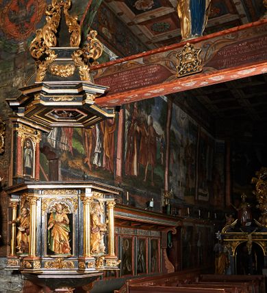 Zdjęcie nr 1: Ambona przyścienna, wisząca, ustawiona na północnym węgarze arkady tęczowej, dostępna przez schody od strony zakrystii. Kosz na planie sześcioboku z wysuniętą w stronę ołtarza częścią prowadzącą do drzwi. Ścianki wydzielone kolumnami o kapitelach korynckich, ustawionymi na postumentach, podtrzymującymi parapet z silnie wysuniętym gzymsem. Między kolumnami płytkie nisze zwieńczone konchami z figurami czterech ewangelistów i Chrystusa. Przyłucza, fryz i cokół dekorowany akantem. Kosz u spodu z podwieszoną szyszką. Wąski zaplecek ujęty pilastrami podtrzymującymi belkowanie. W polu obraz ze św. Janem Nepomucenem w profilowanej ramie w kształcie stojącego prostokąta zamkniętego łukiem nadwieszonym. Przyłucza i fryz dekorowany akantem i rozetami. Baldachim na planie sześcioboku, przedzielony belką arkady tęczowej. Podstawę tworzy belkowanie z wydatnym gzymsem, wyłamane w narożach. Kopuła dekorowana spływami akantowymi oraz girlandami, zwieńczona sześciobocznym cokołem, na którym sześć zwężających się kabłąków utworzonych z ażurowego akantu. Pod baldachimem sześcioboczny kaseton z podwieszoną gołębicą Ducha Świętego. Ambona marmoryzowana w odcieniu ciemnoszarym, ornamentyka i detale architektoniczne złocone, srebrzone i czerwone.