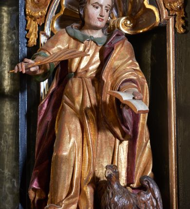 Zdjęcie nr 3: Rzeźba pełnoplastyczna, przedstawiająca św. Jana. Święty zwrócony delikatnie w lewo, ustawiony w kontrapoście z prawą nogą ugiętą w kolanie. Obie ręce ma wyciągnięte na boki, w lewej trzyma otwartą księgę, a w prawej pióro. Twarz młodzieńcza z wysokim czołem, włosy średniej długości z przedziałkiem na środku głowy. Ubrany jest w długą, złotą suknię z zielonym kołnierzem, przepasaną w talii i złoty płaszcz podbity na czerwono, przewieszony przez lewe ramię. Przy lewej nodze orzeł z rozpostartymi skrzydłami. Polichromia naturalistyczna w partiach ciała, szaty złocone.