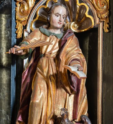 Zdjęcie nr 1: Rzeźba pełnoplastyczna, przedstawiająca św. Jana. Święty zwrócony delikatnie w lewo, ustawiony w kontrapoście z prawą nogą ugiętą w kolanie. Obie ręce ma wyciągnięte na boki, w lewej trzyma otwartą księgę, a w prawej pióro. Twarz młodzieńcza z wysokim czołem, włosy średniej długości z przedziałkiem na środku głowy. Ubrany jest w długą, złotą suknię z zielonym kołnierzem, przepasaną w talii i złoty płaszcz podbity na czerwono, przewieszony przez lewe ramię. Przy lewej nodze orzeł z rozpostartymi skrzydłami. Polichromia naturalistyczna w partiach ciała, szaty złocone.
