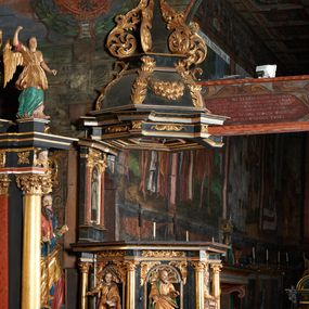 Zdjęcie nr 1: Rzeźba pełnoplastyczna, przedstawiająca św. Łukasza Ewangelistę. Święty ustawiony frontalnie z głową zwróconą w lewo, prawą ręką wyciągniętą przed siebie, w lewej trzyma księgę. Twarz pociągła z wysokim czołem, okolona zarostem. Włosy średniej długości z przedziałkiem na środku głowy, opadające na tył. Ubrany jest w długą, złotą suknię, przepasaną w talii i złoty płaszcz podbity na zielono, przewieszony przez prawe ramię i zawieszony na lewym boku. Przy lewej nodze leży wół. Polichromia naturalistyczna w partiach ciała, szaty złocone.