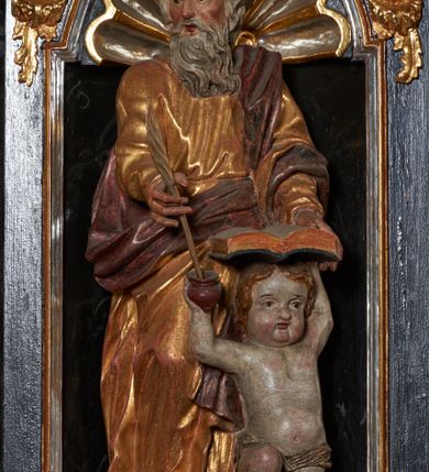 Zdjęcie nr 2: Rzeźba pełnoplastyczna, przedstawiająca św. Mateusza. Figura w delikatnym kontrapoście z prawą nogą ugiętą w kolanie, ustawiona frontalnie, z głową skierowaną w prawą stronę. Lewą ręką podtrzymuje otwartą księgę wspartą na głowie klęczącego u jego lewego boku małego człowieka, zaś prawą trzyma pióro, które macza w kałamarzu znajdującym się w ręku człowieka. Twarz starcza z wysokim czołem, okolona długą i siwą brodą; włosy średniej długości siwe. Ubrany jest w długą, złotą i przepasaną w talii suknię oraz  złoty płaszcz podbity na czerwono, przewieszony przez lewe ramię i zawieszony na prawym boku. Człowiek klęczący na lewym kolanie, z rękami wzniesionymi do góry, w prawej trzyma kałamarz. Twarz pulchna o rysach dziecięcych, włosy krótkie i jasne. Ubrany jest w złoconą tkaninę, przewieszoną przez biodra. Polichromia naturalistyczna w partiach ciała, szaty złocone.