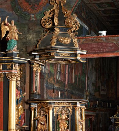 Zdjęcie nr 3: Rzeźba pełnoplastyczna, przedstawiająca św. Mateusza. Figura w delikatnym kontrapoście z prawą nogą ugiętą w kolanie, ustawiona frontalnie, z głową skierowaną w prawą stronę. Lewą ręką podtrzymuje otwartą księgę wspartą na głowie klęczącego u jego lewego boku małego człowieka, zaś prawą trzyma pióro, które macza w kałamarzu znajdującym się w ręku człowieka. Twarz starcza z wysokim czołem, okolona długą i siwą brodą; włosy średniej długości siwe. Ubrany jest w długą, złotą i przepasaną w talii suknię oraz  złoty płaszcz podbity na czerwono, przewieszony przez lewe ramię i zawieszony na prawym boku. Człowiek klęczący na lewym kolanie, z rękami wzniesionymi do góry, w prawej trzyma kałamarz. Twarz pulchna o rysach dziecięcych, włosy krótkie i jasne. Ubrany jest w złoconą tkaninę, przewieszoną przez biodra. Polichromia naturalistyczna w partiach ciała, szaty złocone.