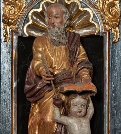 Zdjęcie nr 1: Rzeźba pełnoplastyczna, przedstawiająca św. Mateusza. Figura w delikatnym kontrapoście z prawą nogą ugiętą w kolanie, ustawiona frontalnie, z głową skierowaną w prawą stronę. Lewą ręką podtrzymuje otwartą księgę wspartą na głowie klęczącego u jego lewego boku małego człowieka, zaś prawą trzyma pióro, które macza w kałamarzu znajdującym się w ręku człowieka. Twarz starcza z wysokim czołem, okolona długą i siwą brodą; włosy średniej długości siwe. Ubrany jest w długą, złotą i przepasaną w talii suknię oraz  złoty płaszcz podbity na czerwono, przewieszony przez lewe ramię i zawieszony na prawym boku. Człowiek klęczący na lewym kolanie, z rękami wzniesionymi do góry, w prawej trzyma kałamarz. Twarz pulchna o rysach dziecięcych, włosy krótkie i jasne. Ubrany jest w złoconą tkaninę, przewieszoną przez biodra. Polichromia naturalistyczna w partiach ciała, szaty złocone.