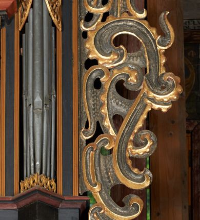 Zdjęcie nr 22: Prospekt organowy podwieszony przy północnej ścianie nawy, oparty na czterech filarach, dostępny z parapetu chóru. Balustrada drewniana z malowaną dekoracją; u spodu pas z ornamentem roślinnym; od frontu podzielona na trzynaście jednofiguralnych przedstawień w ramach, w formie wydłużonych, stojących prostokątów z przedstawieniami aniołów grających na instrumentach. Z boku na balustradzie dwie sceny w formie leżącego prostokąta ze św. Dawidem grającym na lirze i św. Cecylią grająca na organach, oddzielone pasem z dekoracją roślinną. 
Prospekt organowy architektoniczny, pięcioosiowy o bogatej dekoracji snycerskiej. Organy umieszczone na podstawie w kształcie leżącego prostokąta, dekorowanej dwiema płycinami z uszakami w narożach, z malowanymi przedstawieniami grających na instrumentach aniołów oraz z rzeźbionymi uskrzydlonymi główkami anielskimi w glorii pośrodku. Pod cokołem podwieszona dekoracja malarsko-rzeźbiarska nałożona na balustradę chóru. W centrum kompozycji malarskie przedstawienie trzech aniołów (jeden czyta księgę, a dwaj grają na instrumentach) w wieńcu laurowym. Poniżej, rzeźbiona uskrzydlona główka anielska, a całość ujęta ornamentem małżowinowo-chrząstkowym i zakończona u dołu szyszką. Prostokątna podstawa wspiera niski cokół, na którym ustawiona jest trójwieżyczkowa część główna. Środkowa, wyższa wieżyczka jest o przekroju półkolistym, a boczne kątowe o przekroju trójkątnym. Wieżyczki rozdzielają niższe prostokątne płyciny wypełnione piszczałkami. Pod wieżyczkami bocznymi podwieszone są złocone szyszki. Ażurowa dekoracja rzeźbiarska małżowino-chrząstkowa zdobi uszy organu, jest również umieszczona nad płycinami rozdzielającymi wieżyczki. Zwieńczenie tworzą na osiach bocznych strome szczyty, a pośrodku dwugłowy orzeł, na piersi którego w kartuszu w kształcie serca widnieją sygle „A[LBERTUS] B[INKOWICZ] / P[LEBANUS] O[RAVIENSIS]”. Piszczałki przysłaniają od góry ażurowe, rzeźbione firanki o motywie małżowinowo-chrząstkowym, a u dołu wąski pas dekoracji ornamentalnej z motywem silnie przestylizowanych lilii. Organy polichromowane na czarno, profilowania w kolorze czerwonym, zielonym i pomarańczowym, ornamentyka złocona i srebrzona. 



