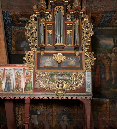 Zdjęcie nr 2: Prospekt organowy podwieszony przy północnej ścianie nawy, oparty na czterech filarach, dostępny z parapetu chóru. Balustrada drewniana z malowaną dekoracją; u spodu pas z ornamentem roślinnym; od frontu podzielona na trzynaście jednofiguralnych przedstawień w ramach, w formie wydłużonych, stojących prostokątów z przedstawieniami aniołów grających na instrumentach. Z boku na balustradzie dwie sceny w formie leżącego prostokąta ze św. Dawidem grającym na lirze i św. Cecylią grająca na organach, oddzielone pasem z dekoracją roślinną. 
Prospekt organowy architektoniczny, pięcioosiowy o bogatej dekoracji snycerskiej. Organy umieszczone na podstawie w kształcie leżącego prostokąta, dekorowanej dwiema płycinami z uszakami w narożach, z malowanymi przedstawieniami grających na instrumentach aniołów oraz z rzeźbionymi uskrzydlonymi główkami anielskimi w glorii pośrodku. Pod cokołem podwieszona dekoracja malarsko-rzeźbiarska nałożona na balustradę chóru. W centrum kompozycji malarskie przedstawienie trzech aniołów (jeden czyta księgę, a dwaj grają na instrumentach) w wieńcu laurowym. Poniżej, rzeźbiona uskrzydlona główka anielska, a całość ujęta ornamentem małżowinowo-chrząstkowym i zakończona u dołu szyszką. Prostokątna podstawa wspiera niski cokół, na którym ustawiona jest trójwieżyczkowa część główna. Środkowa, wyższa wieżyczka jest o przekroju półkolistym, a boczne kątowe o przekroju trójkątnym. Wieżyczki rozdzielają niższe prostokątne płyciny wypełnione piszczałkami. Pod wieżyczkami bocznymi podwieszone są złocone szyszki. Ażurowa dekoracja rzeźbiarska małżowino-chrząstkowa zdobi uszy organu, jest również umieszczona nad płycinami rozdzielającymi wieżyczki. Zwieńczenie tworzą na osiach bocznych strome szczyty, a pośrodku dwugłowy orzeł, na piersi którego w kartuszu w kształcie serca widnieją sygle „A[LBERTUS] B[INKOWICZ] / P[LEBANUS] O[RAVIENSIS]”. Piszczałki przysłaniają od góry ażurowe, rzeźbione firanki o motywie małżowinowo-chrząstkowym, a u dołu wąski pas dekoracji ornamentalnej z motywem silnie przestylizowanych lilii. Organy polichromowane na czarno, profilowania w kolorze czerwonym, zielonym i pomarańczowym, ornamentyka złocona i srebrzona. 



