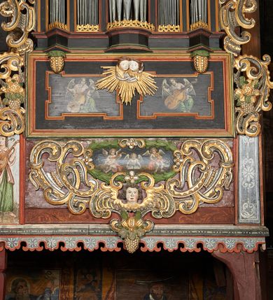 Zdjęcie nr 1: Prospekt organowy podwieszony przy północnej ścianie nawy, oparty na czterech filarach, dostępny z parapetu chóru. Balustrada drewniana z malowaną dekoracją; u spodu pas z ornamentem roślinnym; od frontu podzielona na trzynaście jednofiguralnych przedstawień w ramach, w formie wydłużonych, stojących prostokątów z przedstawieniami aniołów grających na instrumentach. Z boku na balustradzie dwie sceny w formie leżącego prostokąta ze św. Dawidem grającym na lirze i św. Cecylią grająca na organach, oddzielone pasem z dekoracją roślinną. 
Prospekt organowy architektoniczny, pięcioosiowy o bogatej dekoracji snycerskiej. Organy umieszczone na podstawie w kształcie leżącego prostokąta, dekorowanej dwiema płycinami z uszakami w narożach, z malowanymi przedstawieniami grających na instrumentach aniołów oraz z rzeźbionymi uskrzydlonymi główkami anielskimi w glorii pośrodku. Pod cokołem podwieszona dekoracja malarsko-rzeźbiarska nałożona na balustradę chóru. W centrum kompozycji malarskie przedstawienie trzech aniołów (jeden czyta księgę, a dwaj grają na instrumentach) w wieńcu laurowym. Poniżej, rzeźbiona uskrzydlona główka anielska, a całość ujęta ornamentem małżowinowo-chrząstkowym i zakończona u dołu szyszką. Prostokątna podstawa wspiera niski cokół, na którym ustawiona jest trójwieżyczkowa część główna. Środkowa, wyższa wieżyczka jest o przekroju półkolistym, a boczne kątowe o przekroju trójkątnym. Wieżyczki rozdzielają niższe prostokątne płyciny wypełnione piszczałkami. Pod wieżyczkami bocznymi podwieszone są złocone szyszki. Ażurowa dekoracja rzeźbiarska małżowino-chrząstkowa zdobi uszy organu, jest również umieszczona nad płycinami rozdzielającymi wieżyczki. Zwieńczenie tworzą na osiach bocznych strome szczyty, a pośrodku dwugłowy orzeł, na piersi którego w kartuszu w kształcie serca widnieją sygle „A[LBERTUS] B[INKOWICZ] / P[LEBANUS] O[RAVIENSIS]”. Piszczałki przysłaniają od góry ażurowe, rzeźbione firanki o motywie małżowinowo-chrząstkowym, a u dołu wąski pas dekoracji ornamentalnej z motywem silnie przestylizowanych lilii. Organy polichromowane na czarno, profilowania w kolorze czerwonym, zielonym i pomarańczowym, ornamentyka złocona i srebrzona. 



