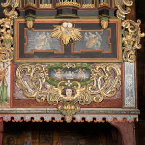 Zdjęcie nr 1: Prospekt organowy podwieszony przy północnej ścianie nawy, oparty na czterech filarach, dostępny z parapetu chóru. Balustrada drewniana z malowaną dekoracją; u spodu pas z ornamentem roślinnym; od frontu podzielona na trzynaście jednofiguralnych przedstawień w ramach, w formie wydłużonych, stojących prostokątów z przedstawieniami aniołów grających na instrumentach. Z boku na balustradzie dwie sceny w formie leżącego prostokąta ze św. Dawidem grającym na lirze i św. Cecylią grająca na organach, oddzielone pasem z dekoracją roślinną. 
Prospekt organowy architektoniczny, pięcioosiowy o bogatej dekoracji snycerskiej. Organy umieszczone na podstawie w kształcie leżącego prostokąta, dekorowanej dwiema płycinami z uszakami w narożach, z malowanymi przedstawieniami grających na instrumentach aniołów oraz z rzeźbionymi uskrzydlonymi główkami anielskimi w glorii pośrodku. Pod cokołem podwieszona dekoracja malarsko-rzeźbiarska nałożona na balustradę chóru. W centrum kompozycji malarskie przedstawienie trzech aniołów (jeden czyta księgę, a dwaj grają na instrumentach) w wieńcu laurowym. Poniżej, rzeźbiona uskrzydlona główka anielska, a całość ujęta ornamentem małżowinowo-chrząstkowym i zakończona u dołu szyszką. Prostokątna podstawa wspiera niski cokół, na którym ustawiona jest trójwieżyczkowa część główna. Środkowa, wyższa wieżyczka jest o przekroju półkolistym, a boczne kątowe o przekroju trójkątnym. Wieżyczki rozdzielają niższe prostokątne płyciny wypełnione piszczałkami. Pod wieżyczkami bocznymi podwieszone są złocone szyszki. Ażurowa dekoracja rzeźbiarska małżowino-chrząstkowa zdobi uszy organu, jest również umieszczona nad płycinami rozdzielającymi wieżyczki. Zwieńczenie tworzą na osiach bocznych strome szczyty, a pośrodku dwugłowy orzeł, na piersi którego w kartuszu w kształcie serca widnieją sygle „A[LBERTUS] B[INKOWICZ] / P[LEBANUS] O[RAVIENSIS]”. Piszczałki przysłaniają od góry ażurowe, rzeźbione firanki o motywie małżowinowo-chrząstkowym, a u dołu wąski pas dekoracji ornamentalnej z motywem silnie przestylizowanych lilii. Organy polichromowane na czarno, profilowania w kolorze czerwonym, zielonym i pomarańczowym, ornamentyka złocona i srebrzona. 



