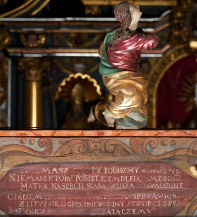 Zdjęcie nr 6: Rzeźba pełna, Chrystus przybity do krzyża trzema gwoździami w lekkim zwisie z głową silnie skręconą w prawo. Twarz podłużna z otwartymi ustami i półprzymkniętymi oczami, okolona kędzierzawą, ciemną brodą; włosy długie spływające na plecy i prawe ramię. Nogi delikatnie ugięte w kolanach; stopy przebite jednym gwoździem, w układzie prawa na lewą. Na głowie Chrystus ma założoną zieloną koronę cierniową. Perizonium w formie tkaniny przewiązanej diagonalnie przez biodra, ze zwisem na prawym boku. Sylwetka wychudzona, smukła, o delikatnym skręcie bioder, z wyraźnie zaznaczonym użyleniem i czerwono-sinymi śladami męki. Polichromia naturalistyczna w partiach ciała, o bladej kolorystyce bieli i błękitu oraz intensywnej czerwonej krwi; perizonium złocone. Nad głową Chrystusa zawieszony zielono-czarny titulus w formie kartusza z napisem „INRI”, ozdobiony motywem małżowinowo-chrząstkowym. Krzyż drewniany, prosty, malowany na czarno. U dołu krzyża zawieszone cztery wota w kształcie złotych serduszek przypiętych czerwonymi kokardkami.