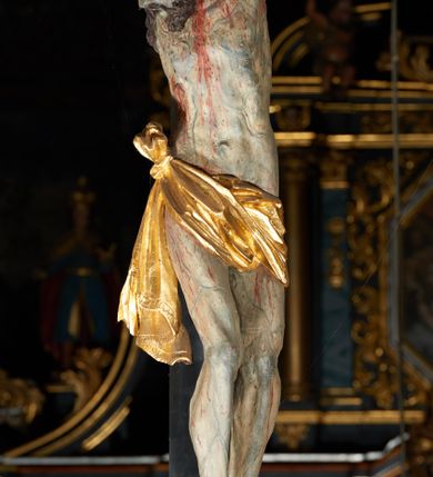 Zdjęcie nr 14: Rzeźba pełna, Chrystus przybity do krzyża trzema gwoździami w lekkim zwisie z głową silnie skręconą w prawo. Twarz podłużna z otwartymi ustami i półprzymkniętymi oczami, okolona kędzierzawą, ciemną brodą; włosy długie spływające na plecy i prawe ramię. Nogi delikatnie ugięte w kolanach; stopy przebite jednym gwoździem, w układzie prawa na lewą. Na głowie Chrystus ma założoną zieloną koronę cierniową. Perizonium w formie tkaniny przewiązanej diagonalnie przez biodra, ze zwisem na prawym boku. Sylwetka wychudzona, smukła, o delikatnym skręcie bioder, z wyraźnie zaznaczonym użyleniem i czerwono-sinymi śladami męki. Polichromia naturalistyczna w partiach ciała, o bladej kolorystyce bieli i błękitu oraz intensywnej czerwonej krwi; perizonium złocone. Nad głową Chrystusa zawieszony zielono-czarny titulus w formie kartusza z napisem „INRI”, ozdobiony motywem małżowinowo-chrząstkowym. Krzyż drewniany, prosty, malowany na czarno. U dołu krzyża zawieszone cztery wota w kształcie złotych serduszek przypiętych czerwonymi kokardkami.