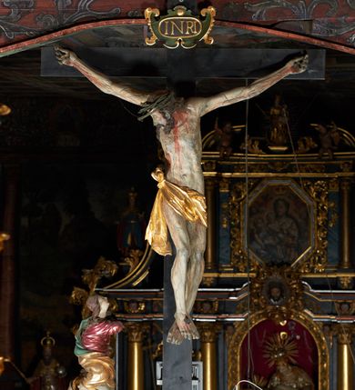 Zdjęcie nr 15: Rzeźba pełna, Chrystus przybity do krzyża trzema gwoździami w lekkim zwisie z głową silnie skręconą w prawo. Twarz podłużna z otwartymi ustami i półprzymkniętymi oczami, okolona kędzierzawą, ciemną brodą; włosy długie spływające na plecy i prawe ramię. Nogi delikatnie ugięte w kolanach; stopy przebite jednym gwoździem, w układzie prawa na lewą. Na głowie Chrystus ma założoną zieloną koronę cierniową. Perizonium w formie tkaniny przewiązanej diagonalnie przez biodra, ze zwisem na prawym boku. Sylwetka wychudzona, smukła, o delikatnym skręcie bioder, z wyraźnie zaznaczonym użyleniem i czerwono-sinymi śladami męki. Polichromia naturalistyczna w partiach ciała, o bladej kolorystyce bieli i błękitu oraz intensywnej czerwonej krwi; perizonium złocone. Nad głową Chrystusa zawieszony zielono-czarny titulus w formie kartusza z napisem „INRI”, ozdobiony motywem małżowinowo-chrząstkowym. Krzyż drewniany, prosty, malowany na czarno. U dołu krzyża zawieszone cztery wota w kształcie złotych serduszek przypiętych czerwonymi kokardkami.