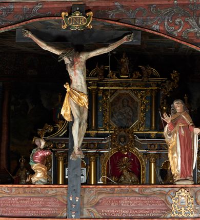 Zdjęcie nr 17: Rzeźba pełna, Chrystus przybity do krzyża trzema gwoździami w lekkim zwisie z głową silnie skręconą w prawo. Twarz podłużna z otwartymi ustami i półprzymkniętymi oczami, okolona kędzierzawą, ciemną brodą; włosy długie spływające na plecy i prawe ramię. Nogi delikatnie ugięte w kolanach; stopy przebite jednym gwoździem, w układzie prawa na lewą. Na głowie Chrystus ma założoną zieloną koronę cierniową. Perizonium w formie tkaniny przewiązanej diagonalnie przez biodra, ze zwisem na prawym boku. Sylwetka wychudzona, smukła, o delikatnym skręcie bioder, z wyraźnie zaznaczonym użyleniem i czerwono-sinymi śladami męki. Polichromia naturalistyczna w partiach ciała, o bladej kolorystyce bieli i błękitu oraz intensywnej czerwonej krwi; perizonium złocone. Nad głową Chrystusa zawieszony zielono-czarny titulus w formie kartusza z napisem „INRI”, ozdobiony motywem małżowinowo-chrząstkowym. Krzyż drewniany, prosty, malowany na czarno. U dołu krzyża zawieszone cztery wota w kształcie złotych serduszek przypiętych czerwonymi kokardkami.