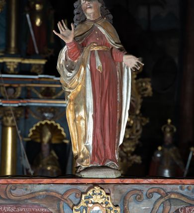 Zdjęcie nr 4: Rzeźba pełna, Chrystus przybity do krzyża trzema gwoździami w lekkim zwisie z głową silnie skręconą w prawo. Twarz podłużna z otwartymi ustami i półprzymkniętymi oczami, okolona kędzierzawą, ciemną brodą; włosy długie spływające na plecy i prawe ramię. Nogi delikatnie ugięte w kolanach; stopy przebite jednym gwoździem, w układzie prawa na lewą. Na głowie Chrystus ma założoną zieloną koronę cierniową. Perizonium w formie tkaniny przewiązanej diagonalnie przez biodra, ze zwisem na prawym boku. Sylwetka wychudzona, smukła, o delikatnym skręcie bioder, z wyraźnie zaznaczonym użyleniem i czerwono-sinymi śladami męki. Polichromia naturalistyczna w partiach ciała, o bladej kolorystyce bieli i błękitu oraz intensywnej czerwonej krwi; perizonium złocone. Nad głową Chrystusa zawieszony zielono-czarny titulus w formie kartusza z napisem „INRI”, ozdobiony motywem małżowinowo-chrząstkowym. Krzyż drewniany, prosty, malowany na czarno. U dołu krzyża zawieszone cztery wota w kształcie złotych serduszek przypiętych czerwonymi kokardkami.