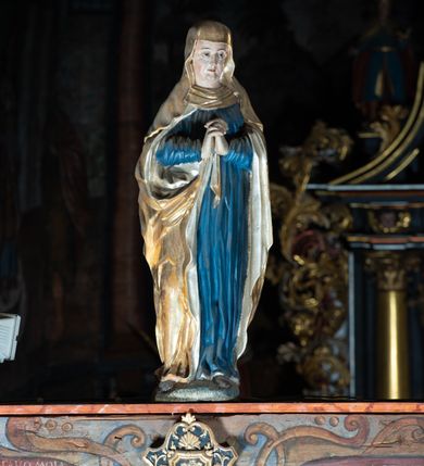 Zdjęcie nr 18: Rzeźba pełna, Chrystus przybity do krzyża trzema gwoździami w lekkim zwisie z głową silnie skręconą w prawo. Twarz podłużna z otwartymi ustami i półprzymkniętymi oczami, okolona kędzierzawą, ciemną brodą; włosy długie spływające na plecy i prawe ramię. Nogi delikatnie ugięte w kolanach; stopy przebite jednym gwoździem, w układzie prawa na lewą. Na głowie Chrystus ma założoną zieloną koronę cierniową. Perizonium w formie tkaniny przewiązanej diagonalnie przez biodra, ze zwisem na prawym boku. Sylwetka wychudzona, smukła, o delikatnym skręcie bioder, z wyraźnie zaznaczonym użyleniem i czerwono-sinymi śladami męki. Polichromia naturalistyczna w partiach ciała, o bladej kolorystyce bieli i błękitu oraz intensywnej czerwonej krwi; perizonium złocone. Nad głową Chrystusa zawieszony zielono-czarny titulus w formie kartusza z napisem „INRI”, ozdobiony motywem małżowinowo-chrząstkowym. Krzyż drewniany, prosty, malowany na czarno. U dołu krzyża zawieszone cztery wota w kształcie złotych serduszek przypiętych czerwonymi kokardkami.