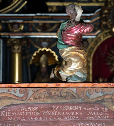 Zdjęcie nr 7: Rzeźba pełna, Chrystus przybity do krzyża trzema gwoździami w lekkim zwisie z głową silnie skręconą w prawo. Twarz podłużna z otwartymi ustami i półprzymkniętymi oczami, okolona kędzierzawą, ciemną brodą; włosy długie spływające na plecy i prawe ramię. Nogi delikatnie ugięte w kolanach; stopy przebite jednym gwoździem, w układzie prawa na lewą. Na głowie Chrystus ma założoną zieloną koronę cierniową. Perizonium w formie tkaniny przewiązanej diagonalnie przez biodra, ze zwisem na prawym boku. Sylwetka wychudzona, smukła, o delikatnym skręcie bioder, z wyraźnie zaznaczonym użyleniem i czerwono-sinymi śladami męki. Polichromia naturalistyczna w partiach ciała, o bladej kolorystyce bieli i błękitu oraz intensywnej czerwonej krwi; perizonium złocone. Nad głową Chrystusa zawieszony zielono-czarny titulus w formie kartusza z napisem „INRI”, ozdobiony motywem małżowinowo-chrząstkowym. Krzyż drewniany, prosty, malowany na czarno. U dołu krzyża zawieszone cztery wota w kształcie złotych serduszek przypiętych czerwonymi kokardkami.