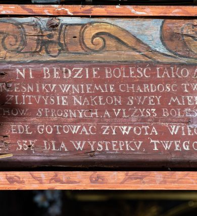 Zdjęcie nr 9: Rzeźba pełna, Chrystus przybity do krzyża trzema gwoździami w lekkim zwisie z głową silnie skręconą w prawo. Twarz podłużna z otwartymi ustami i półprzymkniętymi oczami, okolona kędzierzawą, ciemną brodą; włosy długie spływające na plecy i prawe ramię. Nogi delikatnie ugięte w kolanach; stopy przebite jednym gwoździem, w układzie prawa na lewą. Na głowie Chrystus ma założoną zieloną koronę cierniową. Perizonium w formie tkaniny przewiązanej diagonalnie przez biodra, ze zwisem na prawym boku. Sylwetka wychudzona, smukła, o delikatnym skręcie bioder, z wyraźnie zaznaczonym użyleniem i czerwono-sinymi śladami męki. Polichromia naturalistyczna w partiach ciała, o bladej kolorystyce bieli i błękitu oraz intensywnej czerwonej krwi; perizonium złocone. Nad głową Chrystusa zawieszony zielono-czarny titulus w formie kartusza z napisem „INRI”, ozdobiony motywem małżowinowo-chrząstkowym. Krzyż drewniany, prosty, malowany na czarno. U dołu krzyża zawieszone cztery wota w kształcie złotych serduszek przypiętych czerwonymi kokardkami.