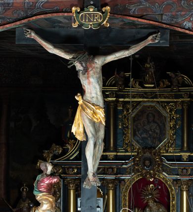 Zdjęcie nr 1: Rzeźba pełna, Chrystus przybity do krzyża trzema gwoździami w lekkim zwisie z głową silnie skręconą w prawo. Twarz podłużna z otwartymi ustami i półprzymkniętymi oczami, okolona kędzierzawą, ciemną brodą; włosy długie spływające na plecy i prawe ramię. Nogi delikatnie ugięte w kolanach; stopy przebite jednym gwoździem, w układzie prawa na lewą. Na głowie Chrystus ma założoną zieloną koronę cierniową. Perizonium w formie tkaniny przewiązanej diagonalnie przez biodra, ze zwisem na prawym boku. Sylwetka wychudzona, smukła, o delikatnym skręcie bioder, z wyraźnie zaznaczonym użyleniem i czerwono-sinymi śladami męki. Polichromia naturalistyczna w partiach ciała, o bladej kolorystyce bieli i błękitu oraz intensywnej czerwonej krwi; perizonium złocone. Nad głową Chrystusa zawieszony zielono-czarny titulus w formie kartusza z napisem „INRI”, ozdobiony motywem małżowinowo-chrząstkowym. Krzyż drewniany, prosty, malowany na czarno. U dołu krzyża zawieszone cztery wota w kształcie złotych serduszek przypiętych czerwonymi kokardkami.