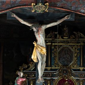 Zdjęcie nr 1: Rzeźba pełna, Chrystus przybity do krzyża trzema gwoździami w lekkim zwisie z głową silnie skręconą w prawo. Twarz podłużna z otwartymi ustami i półprzymkniętymi oczami, okolona kędzierzawą, ciemną brodą; włosy długie spływające na plecy i prawe ramię. Nogi delikatnie ugięte w kolanach; stopy przebite jednym gwoździem, w układzie prawa na lewą. Na głowie Chrystus ma założoną zieloną koronę cierniową. Perizonium w formie tkaniny przewiązanej diagonalnie przez biodra, ze zwisem na prawym boku. Sylwetka wychudzona, smukła, o delikatnym skręcie bioder, z wyraźnie zaznaczonym użyleniem i czerwono-sinymi śladami męki. Polichromia naturalistyczna w partiach ciała, o bladej kolorystyce bieli i błękitu oraz intensywnej czerwonej krwi; perizonium złocone. Nad głową Chrystusa zawieszony zielono-czarny titulus w formie kartusza z napisem „INRI”, ozdobiony motywem małżowinowo-chrząstkowym. Krzyż drewniany, prosty, malowany na czarno. U dołu krzyża zawieszone cztery wota w kształcie złotych serduszek przypiętych czerwonymi kokardkami.