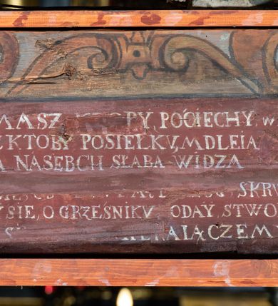 Zdjęcie nr 10: Rzeźba pełna, Chrystus przybity do krzyża trzema gwoździami w lekkim zwisie z głową silnie skręconą w prawo. Twarz podłużna z otwartymi ustami i półprzymkniętymi oczami, okolona kędzierzawą, ciemną brodą; włosy długie spływające na plecy i prawe ramię. Nogi delikatnie ugięte w kolanach; stopy przebite jednym gwoździem, w układzie prawa na lewą. Na głowie Chrystus ma założoną zieloną koronę cierniową. Perizonium w formie tkaniny przewiązanej diagonalnie przez biodra, ze zwisem na prawym boku. Sylwetka wychudzona, smukła, o delikatnym skręcie bioder, z wyraźnie zaznaczonym użyleniem i czerwono-sinymi śladami męki. Polichromia naturalistyczna w partiach ciała, o bladej kolorystyce bieli i błękitu oraz intensywnej czerwonej krwi; perizonium złocone. Nad głową Chrystusa zawieszony zielono-czarny titulus w formie kartusza z napisem „INRI”, ozdobiony motywem małżowinowo-chrząstkowym. Krzyż drewniany, prosty, malowany na czarno. U dołu krzyża zawieszone cztery wota w kształcie złotych serduszek przypiętych czerwonymi kokardkami.